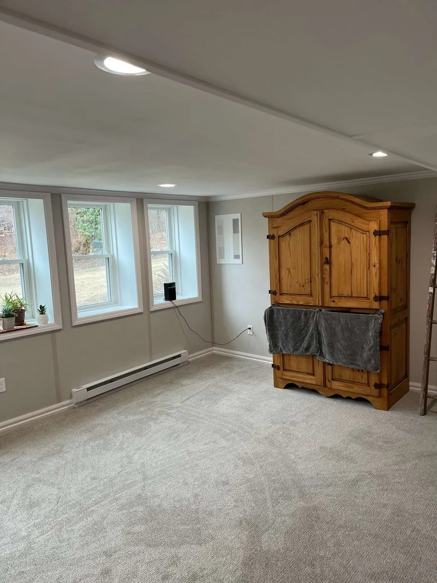 Bright basement room with light gray walls and carpet. Large wooden armoire with a gray towel hangs against the wall. Three windows display an outside view.