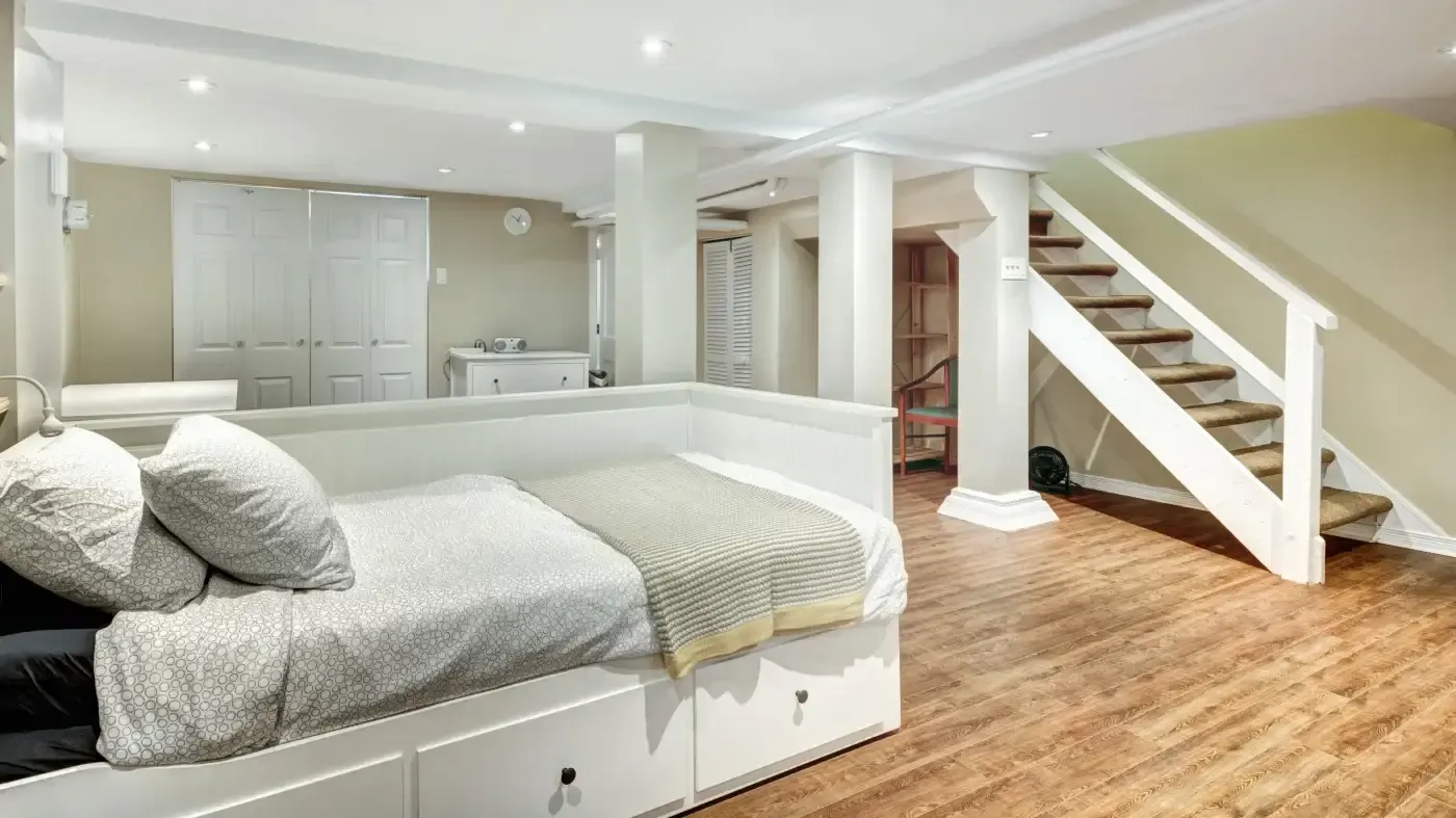A cozy basement room features a bed with gray bedding, white storage drawers, wooden flooring, and a staircase. The scene is well-lit, conveying a warm, inviting tone.
