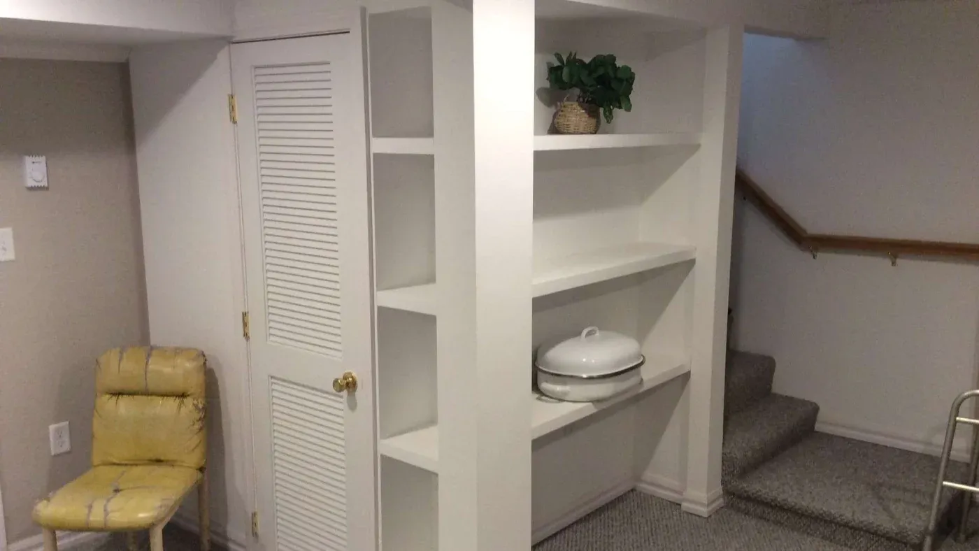 A basement with white built-in shelves, holding a potted plant and a white dish. Nearby, a worn yellow chair and a carpeted staircase are visible.