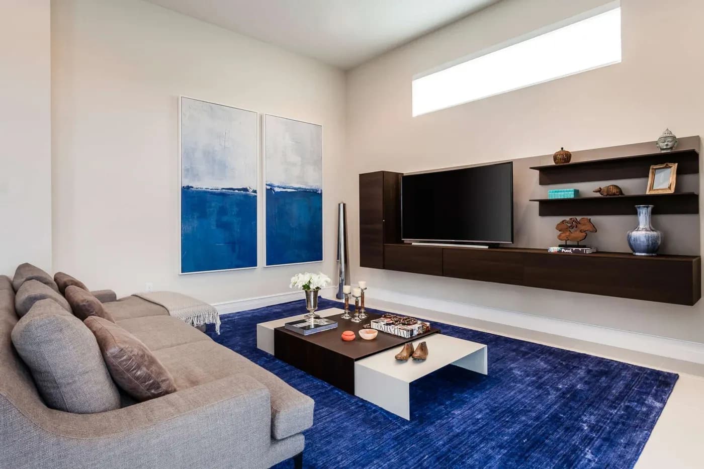 Modern living room with a gray sofa, large blue abstract paintings, and a sleek wall-mounted TV. A blue rug complements dark wood furniture, creating a cozy feel.