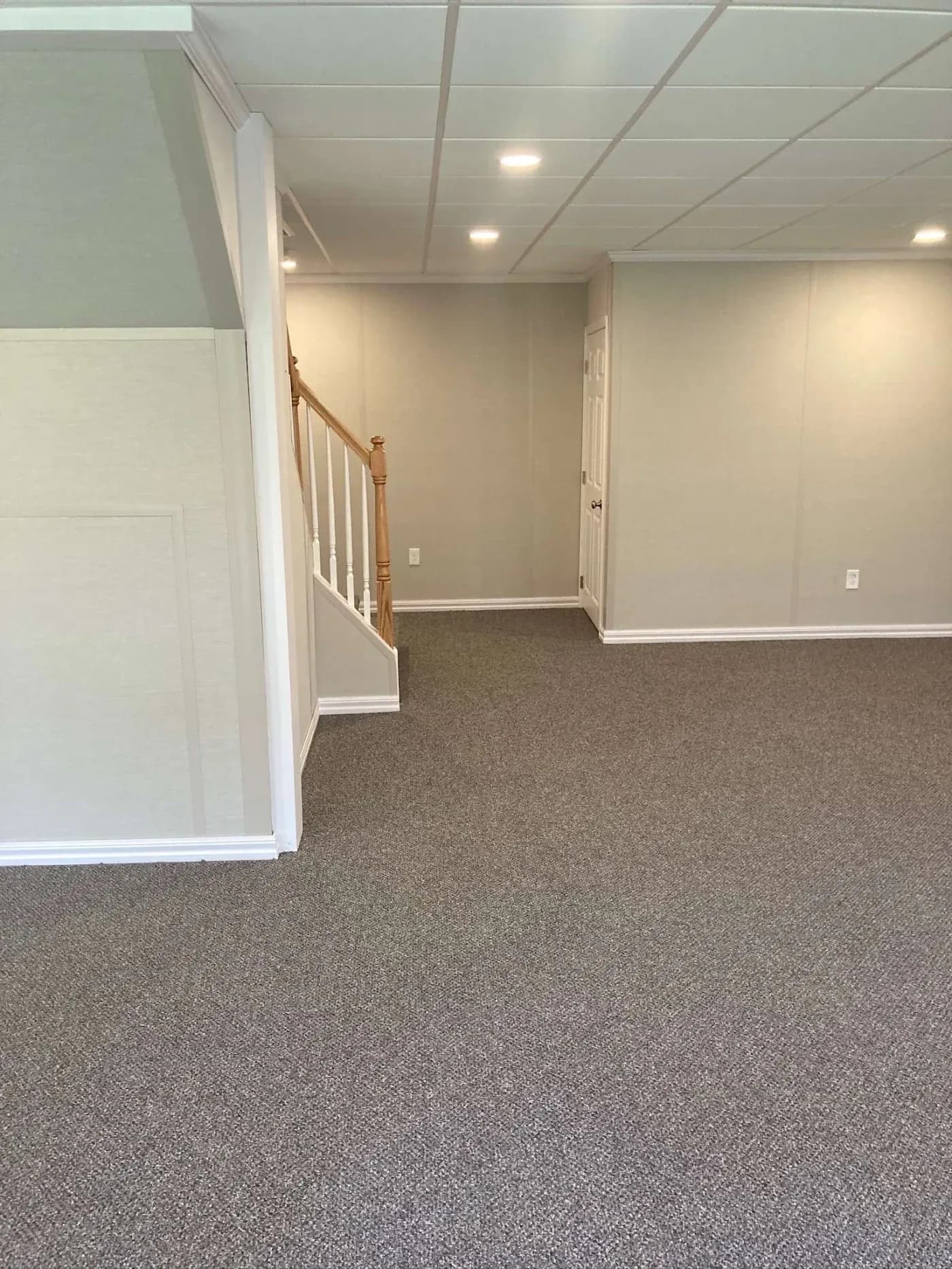 Modern basement with gray carpet, white walls, and a ceiling with recessed lights. Wooden staircase leads up, creating a minimalist, spacious feel.