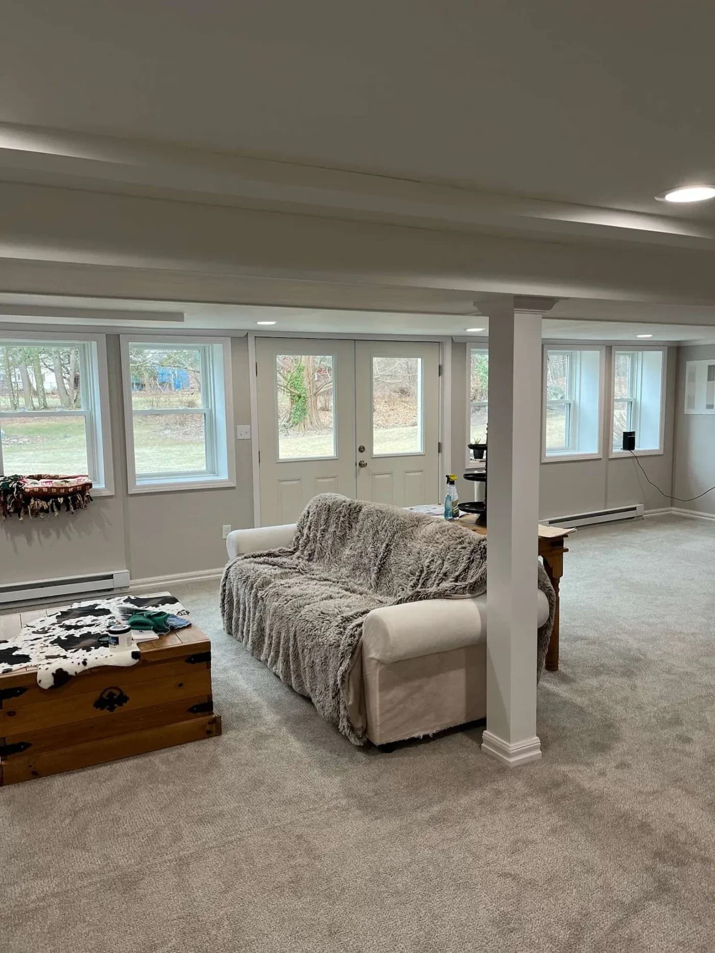A cozy basement features a beige sofa with a furry throw, facing a wooden chest used as a coffee table. Natural light streams through multiple windows, illuminating the carpeted space and creating a calm, inviting atmosphere.