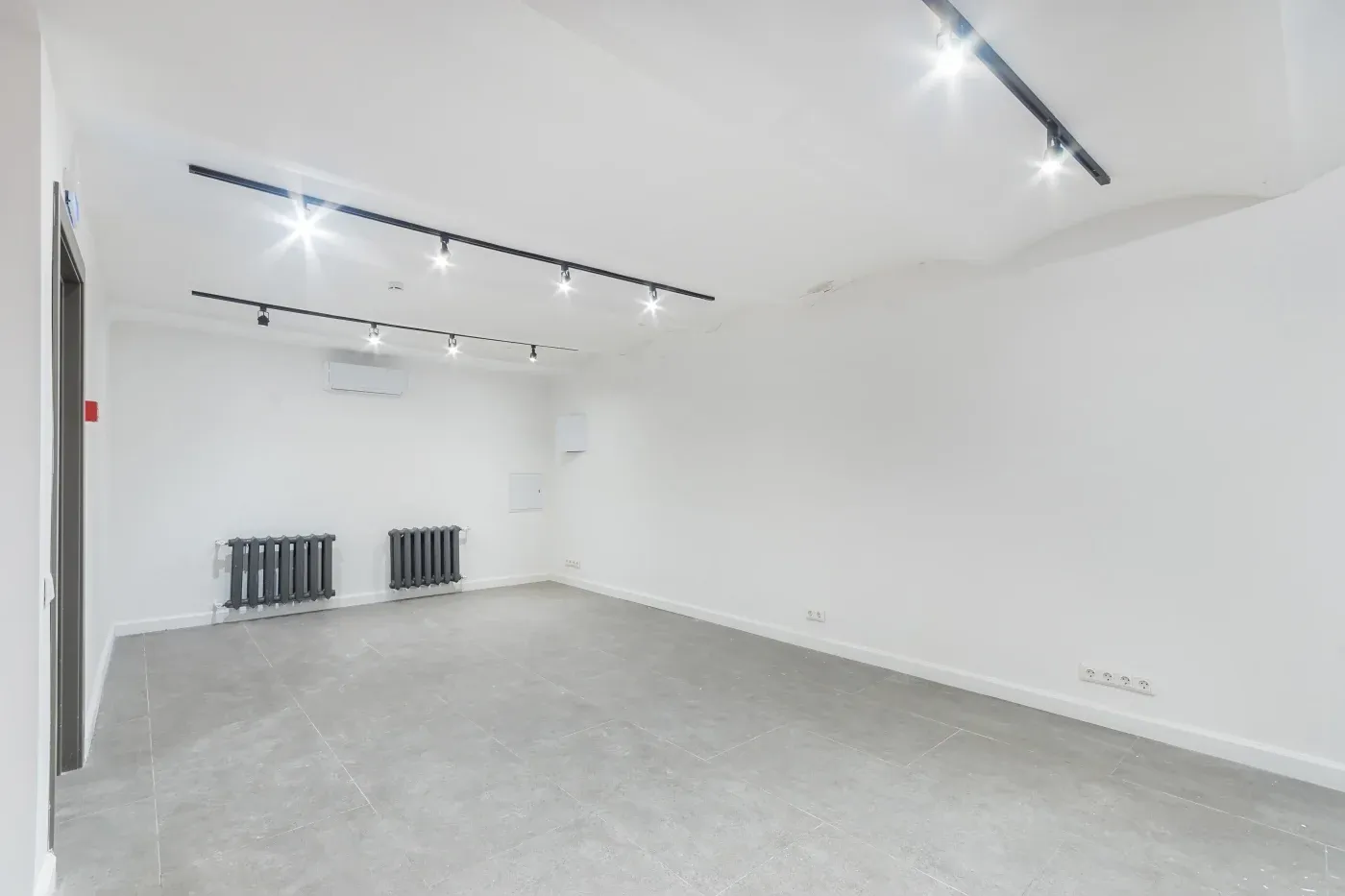 Spacious, empty white room with gray tiled floor, bright track lighting on the ceiling, a black radiator, an air vent, and minimal decor.