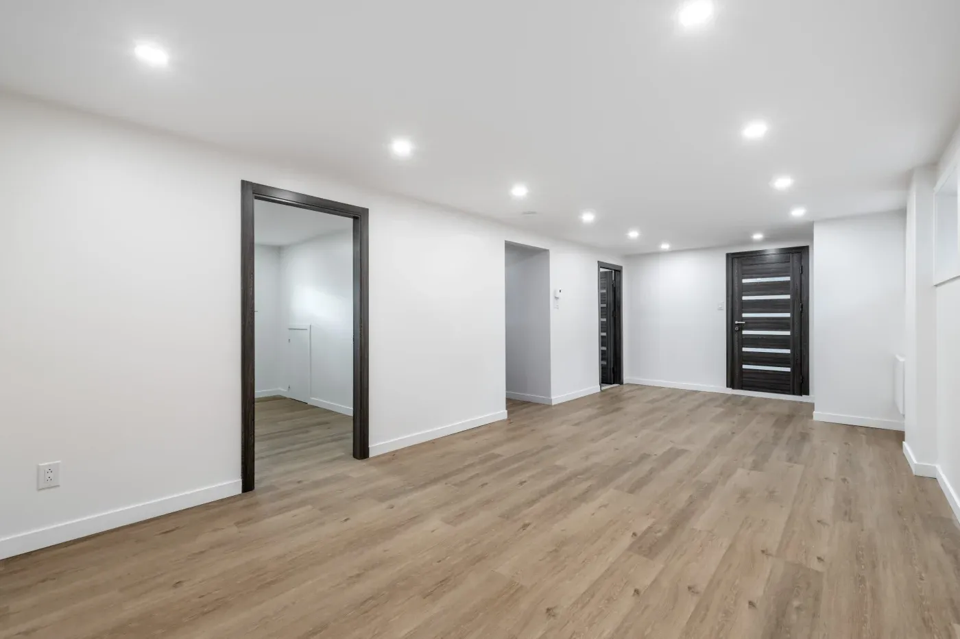 Spacious empty room with light wood flooring, white walls, and recessed ceiling lights. Dark wooden doors add contrast, creating a modern, clean look.