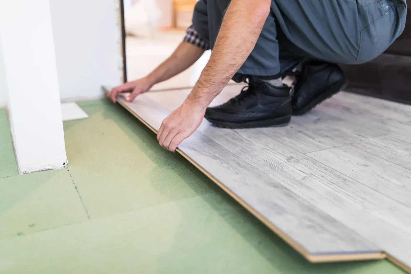 A person installs light gray laminate flooring, kneeling with hands firmly pressing the plank into place. The scene conveys careful precision and focus.