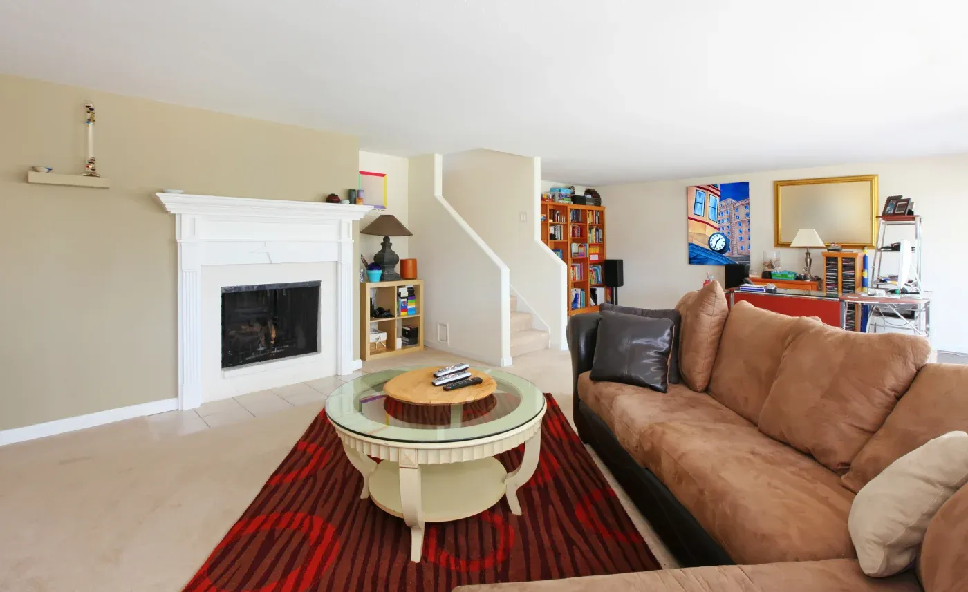 A cozy living room with beige walls features a brown sofa, a round glass coffee table on a red rug, a white fireplace, and a bookshelf by the staircase.