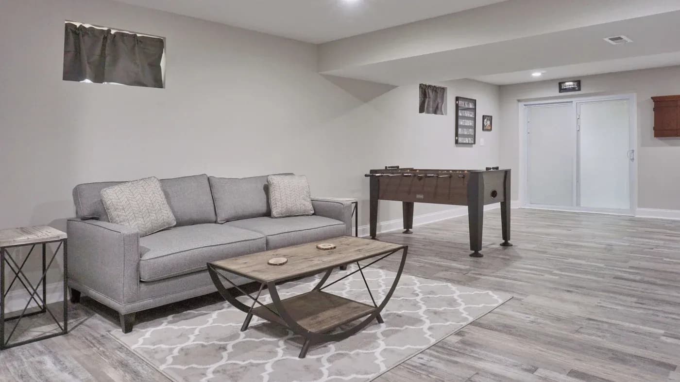 Modern basement with a gray sofa, patterned rug, and wooden coffee table, creating a cozy ambiance. A foosball table adds a playful touch.