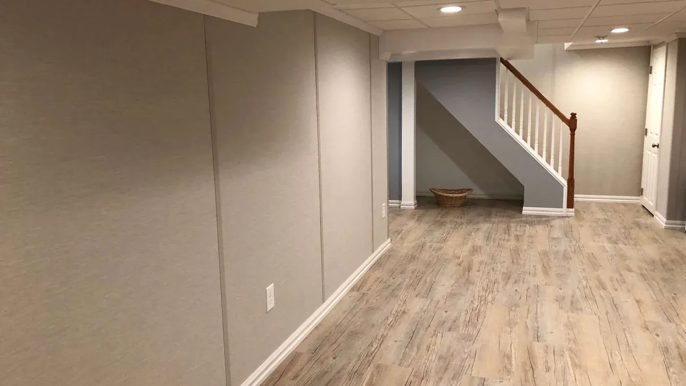 A spacious, empty basement with light wood flooring and beige walls. Recessed lighting brightens the room, and a wooden staircase leads upward, adding depth.