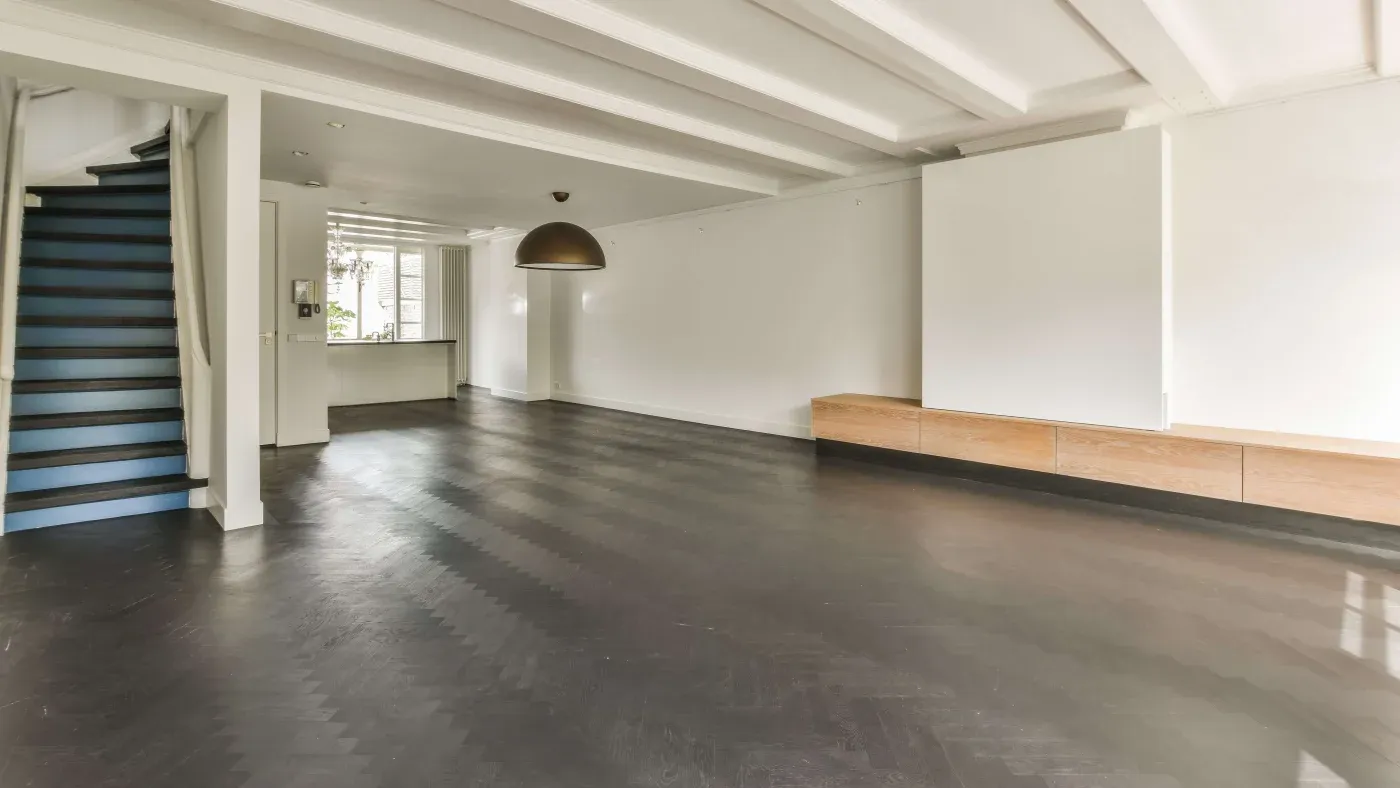 Spacious empty room with dark hardwood floors, white walls, and a coffered ceiling. Features a staircase, large windows, and a modern pendant light.
