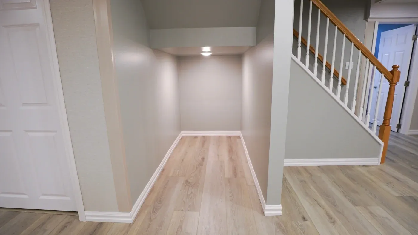A small, well-lit alcove with light wood flooring, located under a staircase. The walls are painted light gray, evoking a calm and cozy atmosphere.