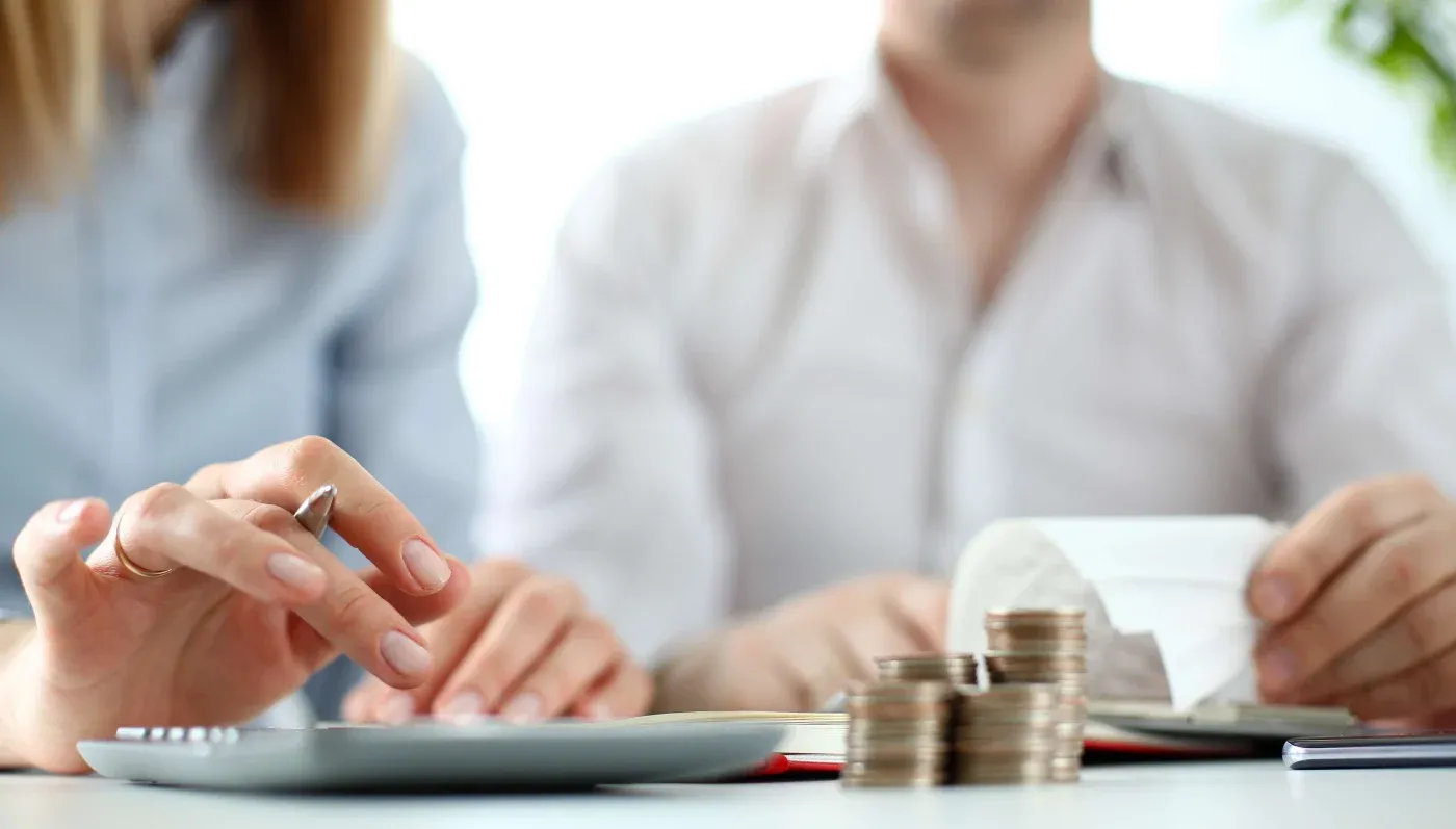 Two people working at a desk; one uses a calculator while the other holds a receipt. Stacks of coins are in the foreground, suggesting budgeting or finance.