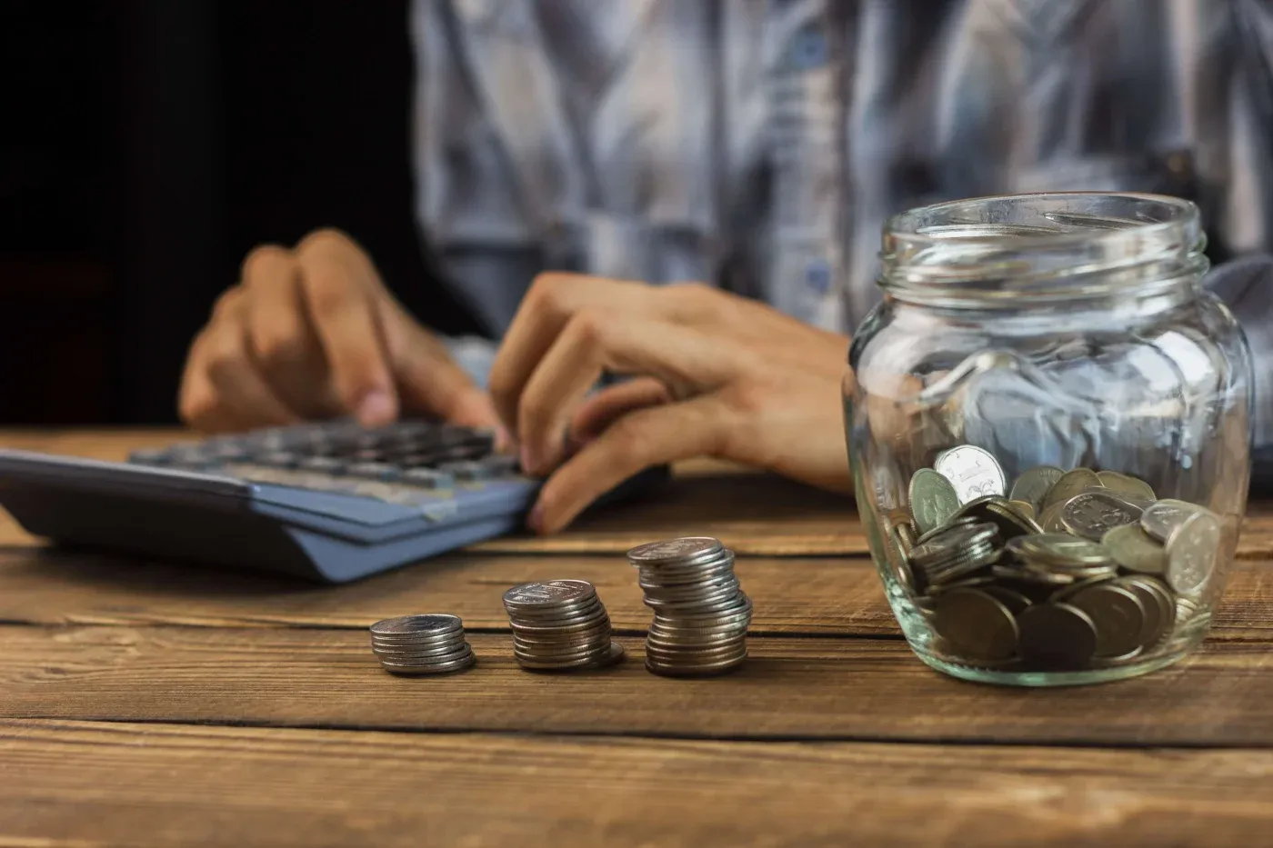 A person wearing a plaid shirt uses a calculator behind stacked coins and a jar of coins on a wooden table, conveying a budgeting or financial planning theme.