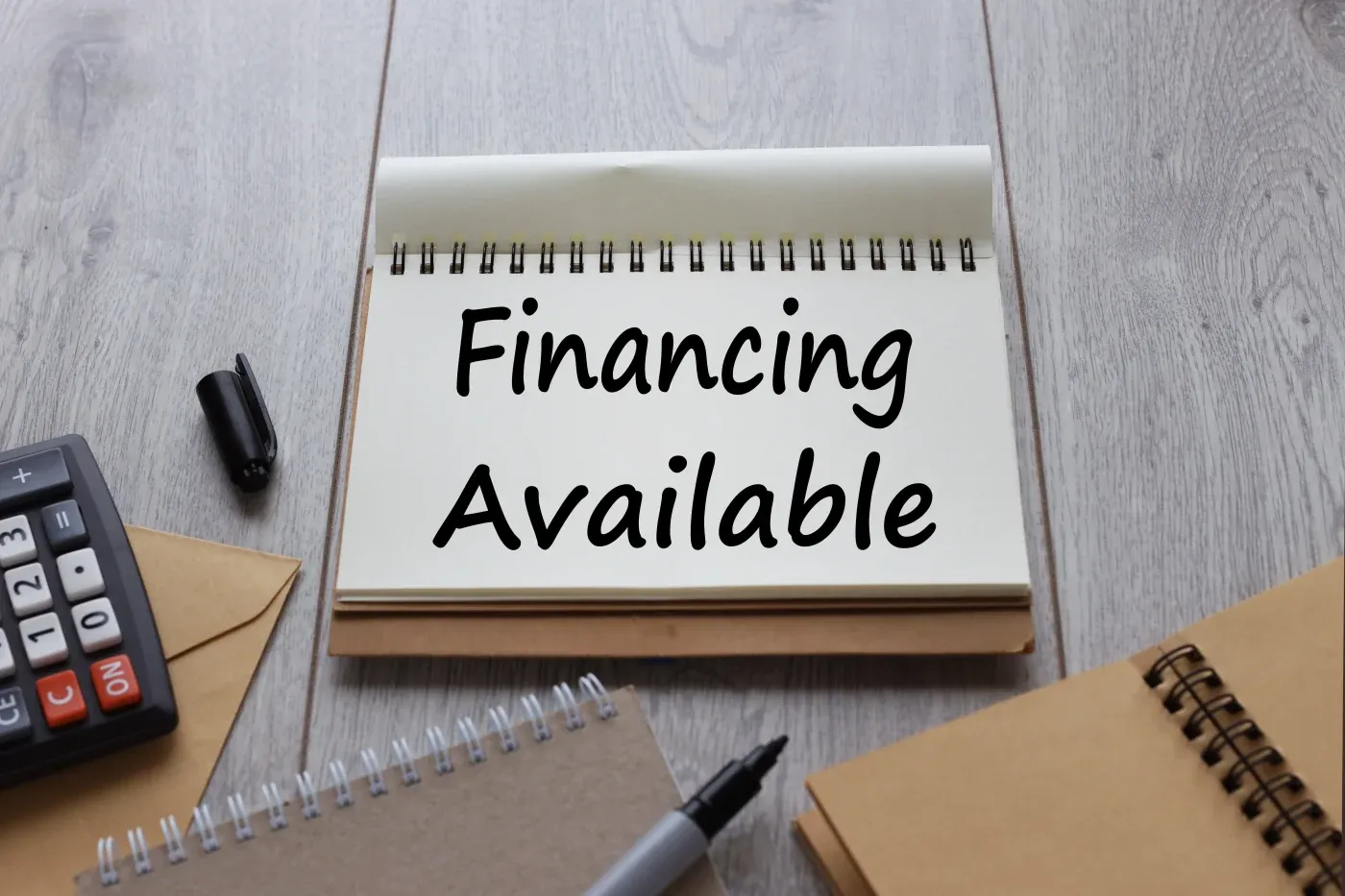 An open notebook on a wooden table reads "Financing Available." Surrounding items include a pen, a calculator, and two brown notebooks, suggesting business planning.