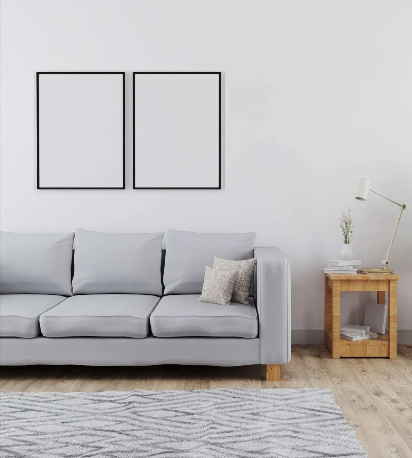 Minimalist living room with a light gray sofa, two blank frames on a white wall, a wooden side table with a lamp, and a patterned rug on wooden floor.