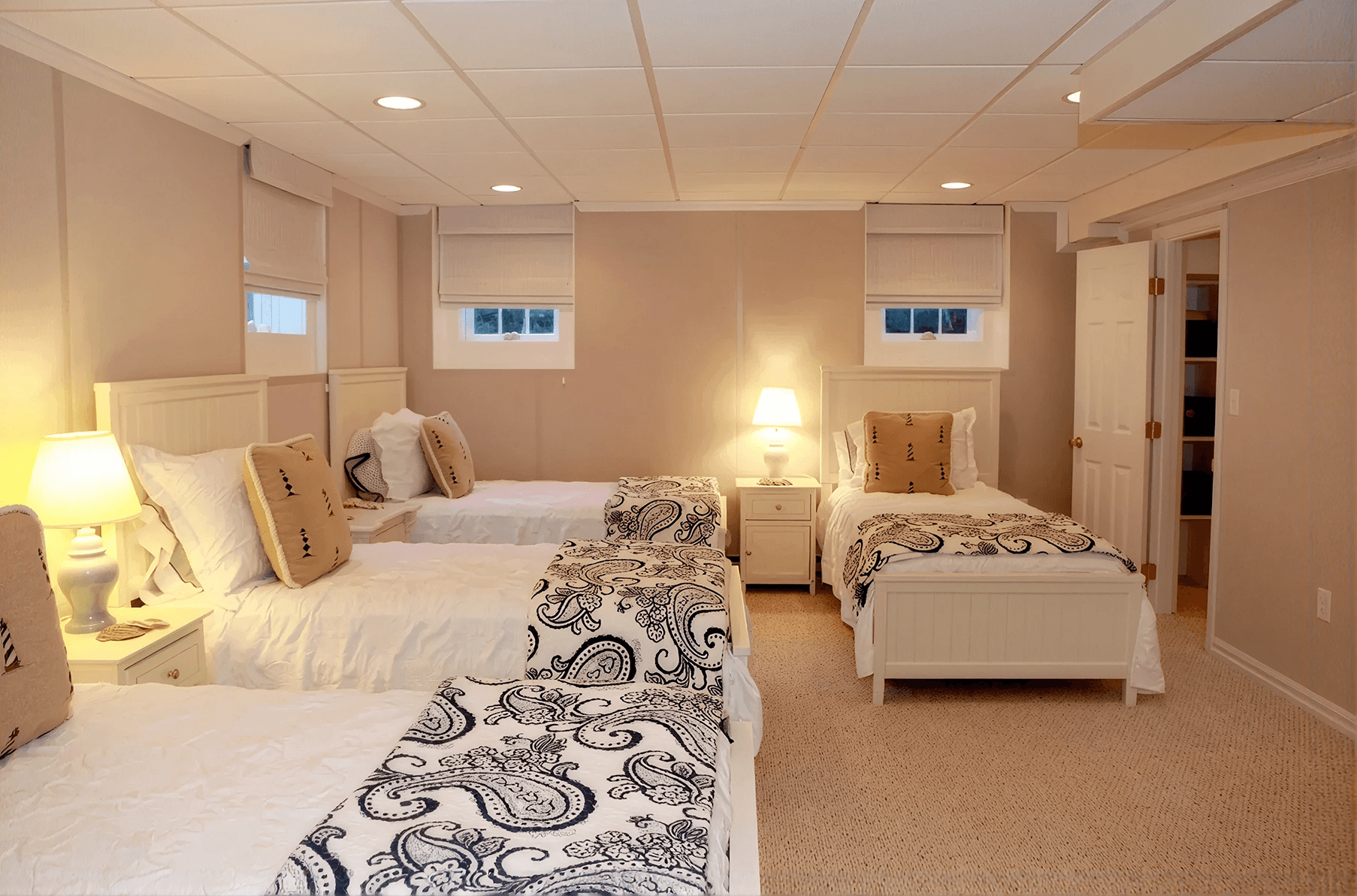 Finished basement bedroom with four white twin beds, matching nightstands, soft carpeting, and neutral walls. Small windows, recessed lighting, and coordinated bedding create a bright, comfortable space for accommodating multiple guests.