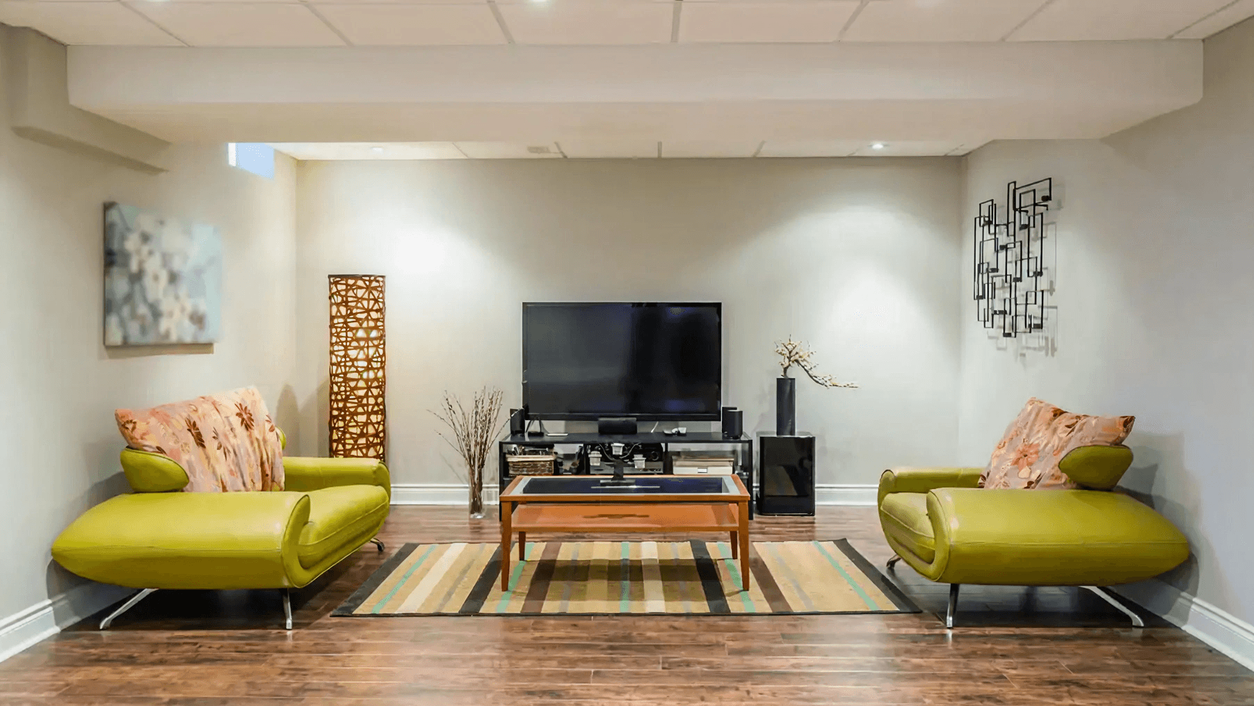 Modern living room with a cozy vibe, featuring two green sofas with floral cushions, a flat-screen TV, wooden coffee table, and decorative art pieces on a neutral wall.