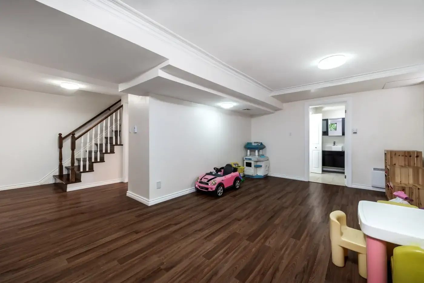 Spacious basement playroom with wooden floors, pink toy car, kitchen set, and colorful table. Stairs to the left and a doorway to the right. Bright, welcoming atmosphere.