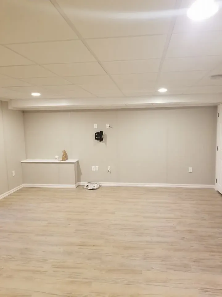 A minimalist basement room with beige walls and light wood flooring. It features a wall-mounted bracket, a recessed shelf, and ceiling lights, conveying calmness.