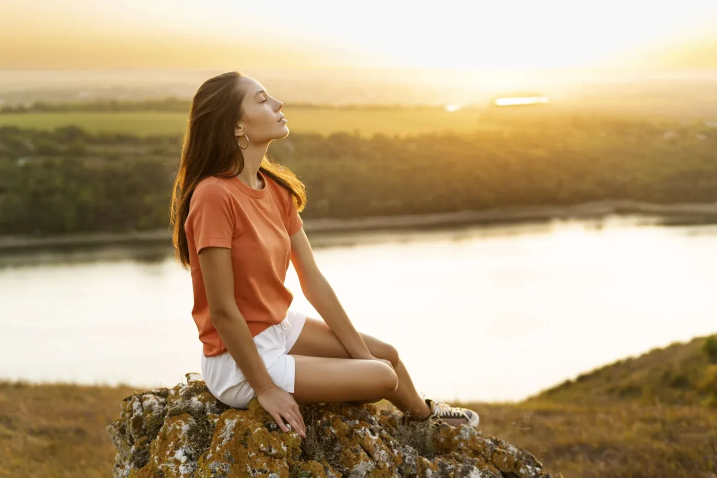 A woman in an orange shirt and white shorts sits peacefully on a rocky hilltop, eyes closed, at sunset overlooking a serene river landscape.