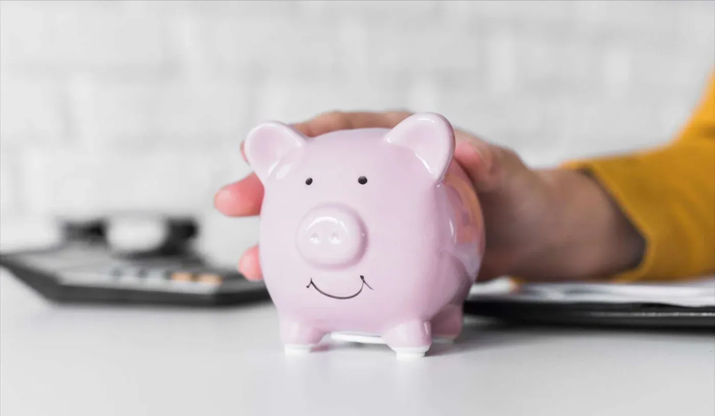 A person in a yellow sleeve gently holds a pink piggy bank on a white table, with a blurred calculator in the background. The scene conveys saving money.