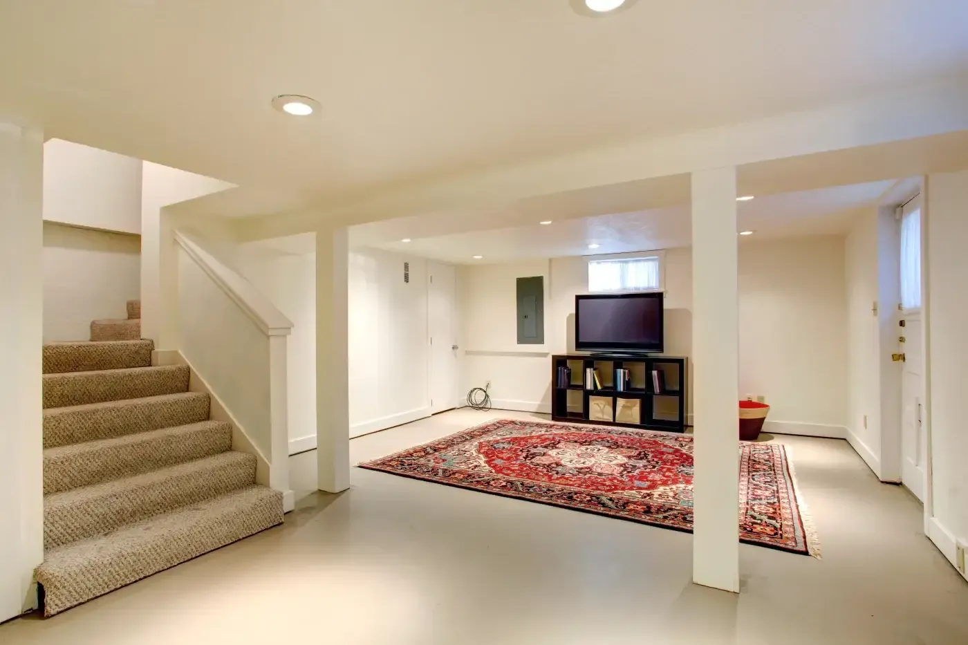 Basement room with beige walls and carpeted stairs. Features a colorful patterned rug, TV on a stand, bookshelves, and a cozy, well-lit atmosphere.
