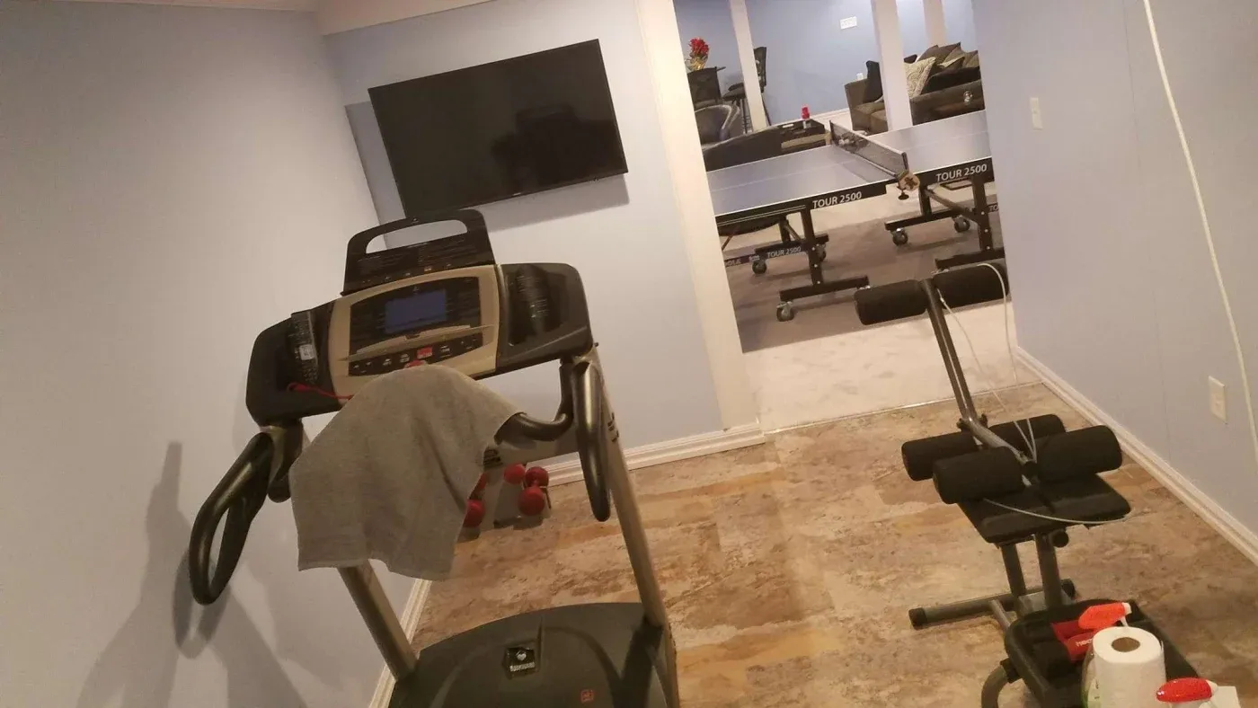 Home gym featuring a treadmill with a towel, small weights, an exercise bench, a wall-mounted TV, and a nearby ping pong table in a cozy setting.