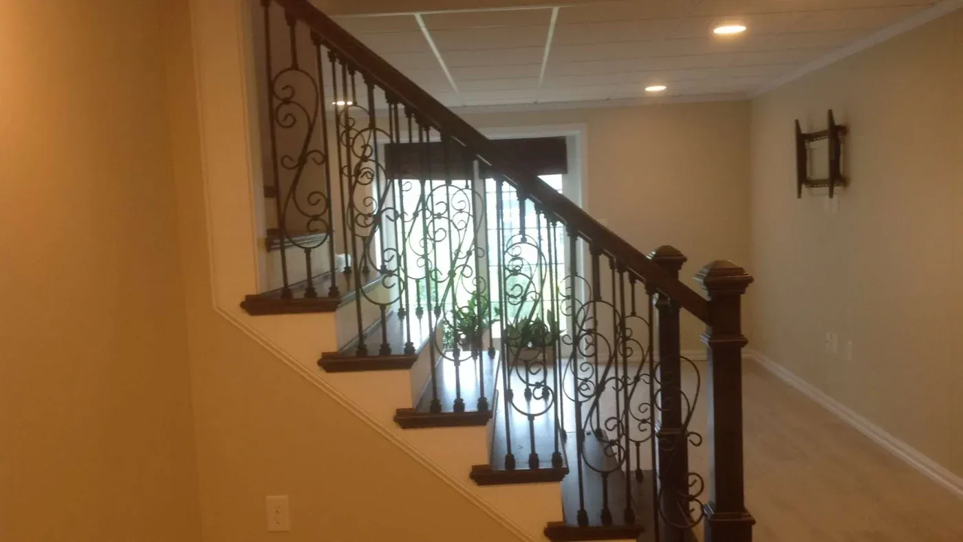 A staircase with ornate wrought iron railings in a warmly lit hallway. Light wood floors and a potted plant near a large window create a cozy ambiance.