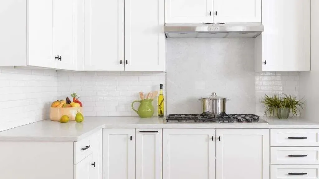 Bright kitchen with white cabinets, sleek metal stove, and a pot. Green vase, fruit bowl, oil bottle, and plant add color. Clean, modern vibe.