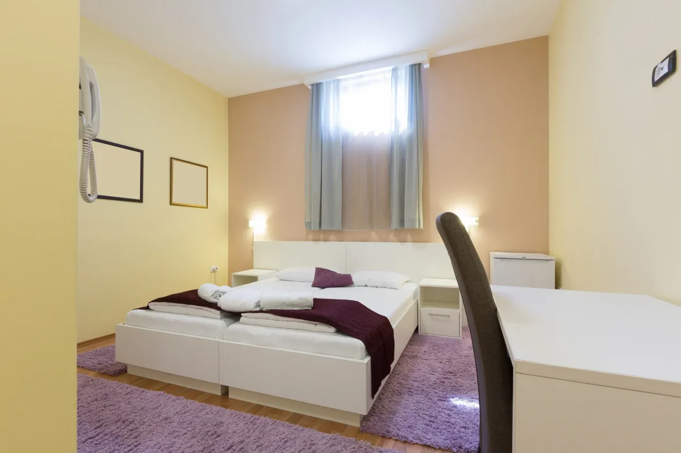 Modern hotel room with a double bed, white linens, and purple accents. Soft lighting, wall art, a desk, and window with sheer curtains create a cozy ambiance.