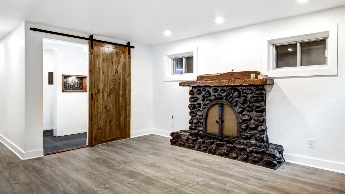 Rustic living room with a stone fireplace and wooden mantel. A sliding barn door leads to a tiled space. The room has wooden floors and white walls, creating a cozy ambiance.