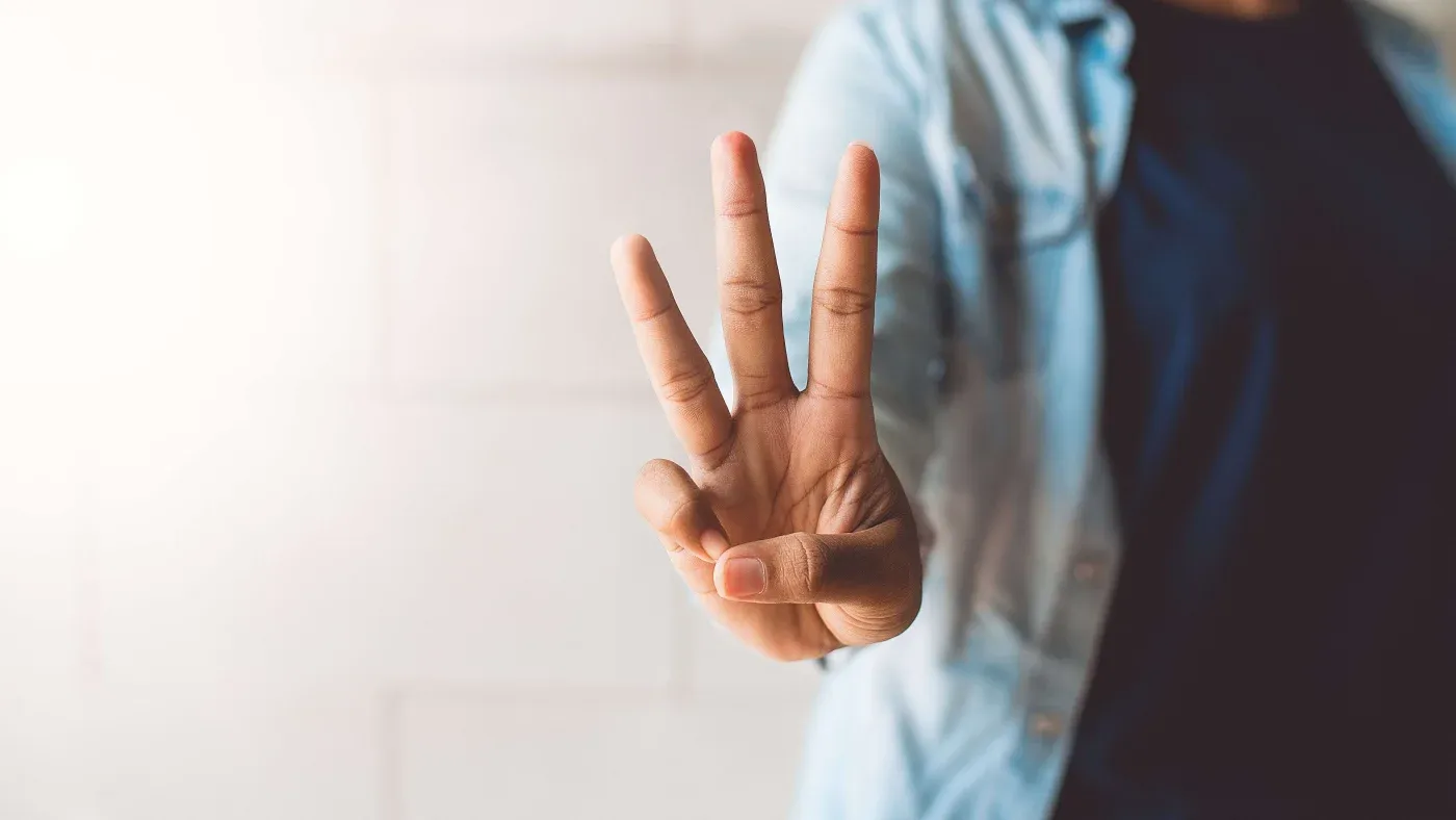 A person holds up three fingers against a softly lit background, wearing a blue shirt, conveying a sense of calm and simplicity.