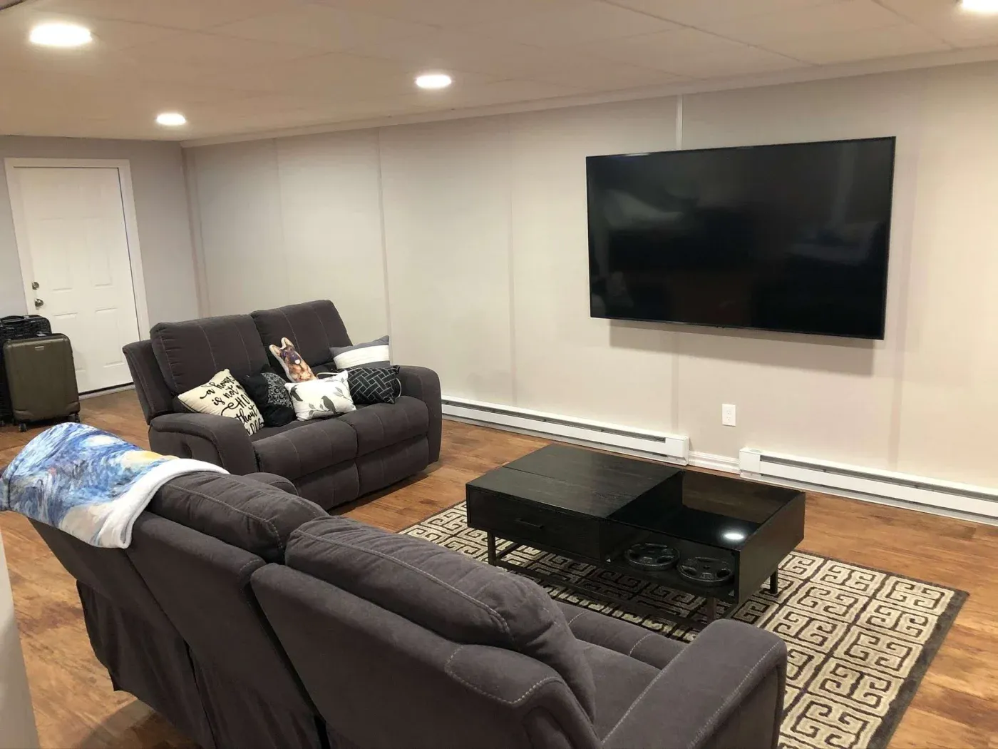 A cozy living room with gray sofas adorned with patterned cushions and throws. A large flat-screen TV is mounted on the opposite wall. A dark coffee table sits on a geometric rug, conveying a modern and inviting atmosphere.