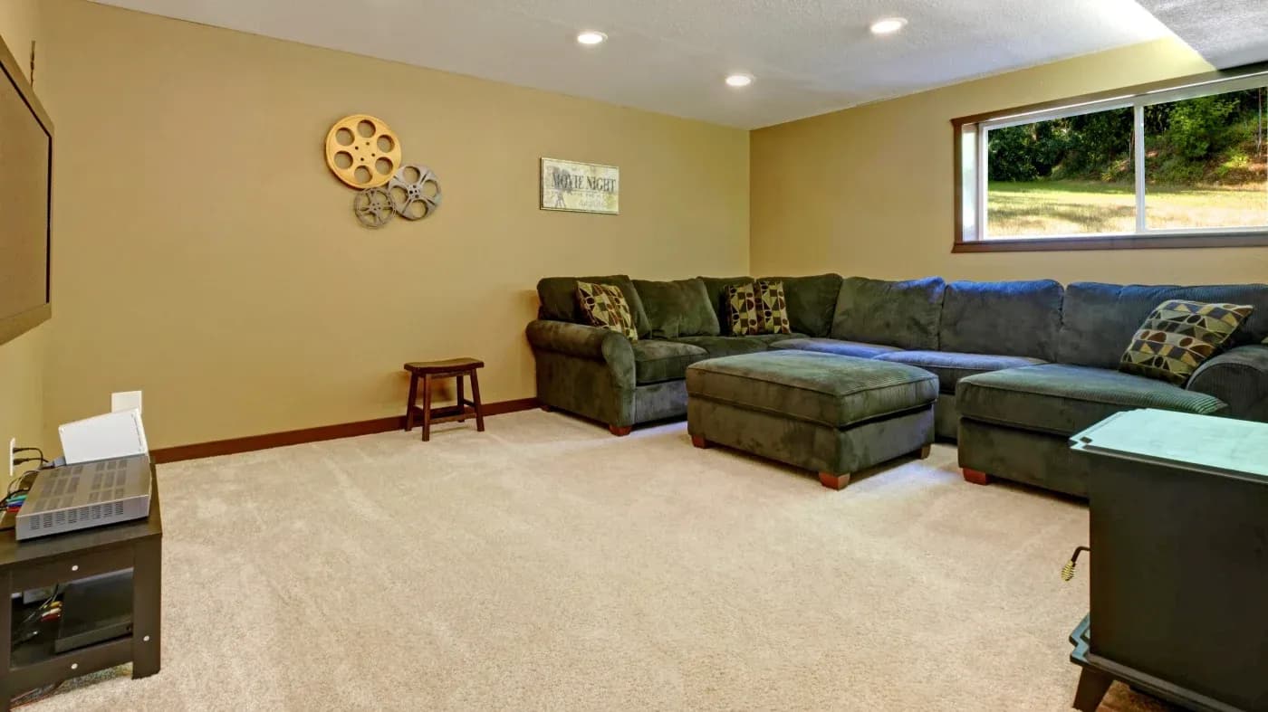 Cozy basement family room with a large sectional sofa, ottoman, film-reel wall decor, soft carpet, and a bright window overlooking greenery.
