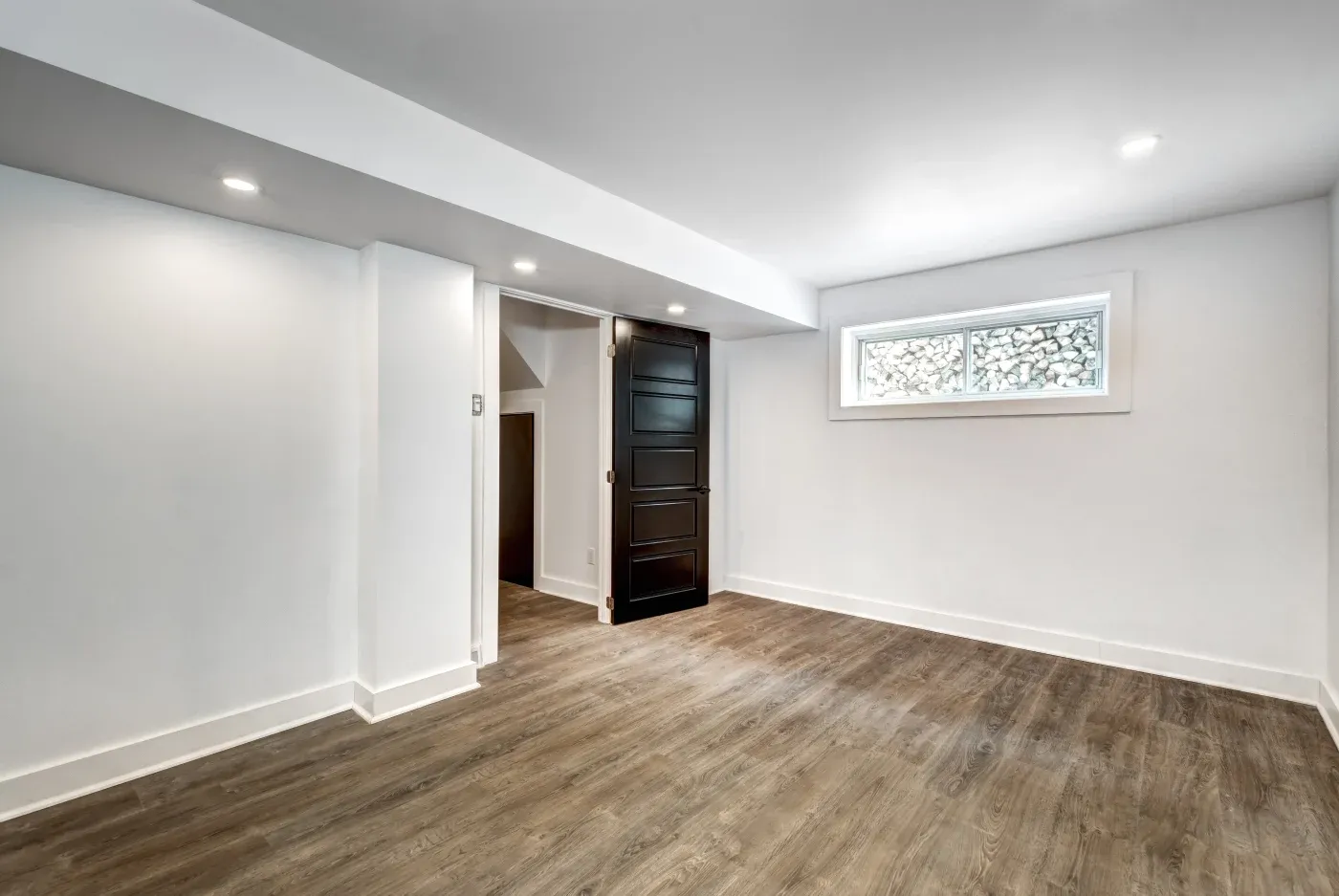 Spacious basement room with white walls, wood floor, small high window, and an open black door. Recessed lighting adds brightness.
