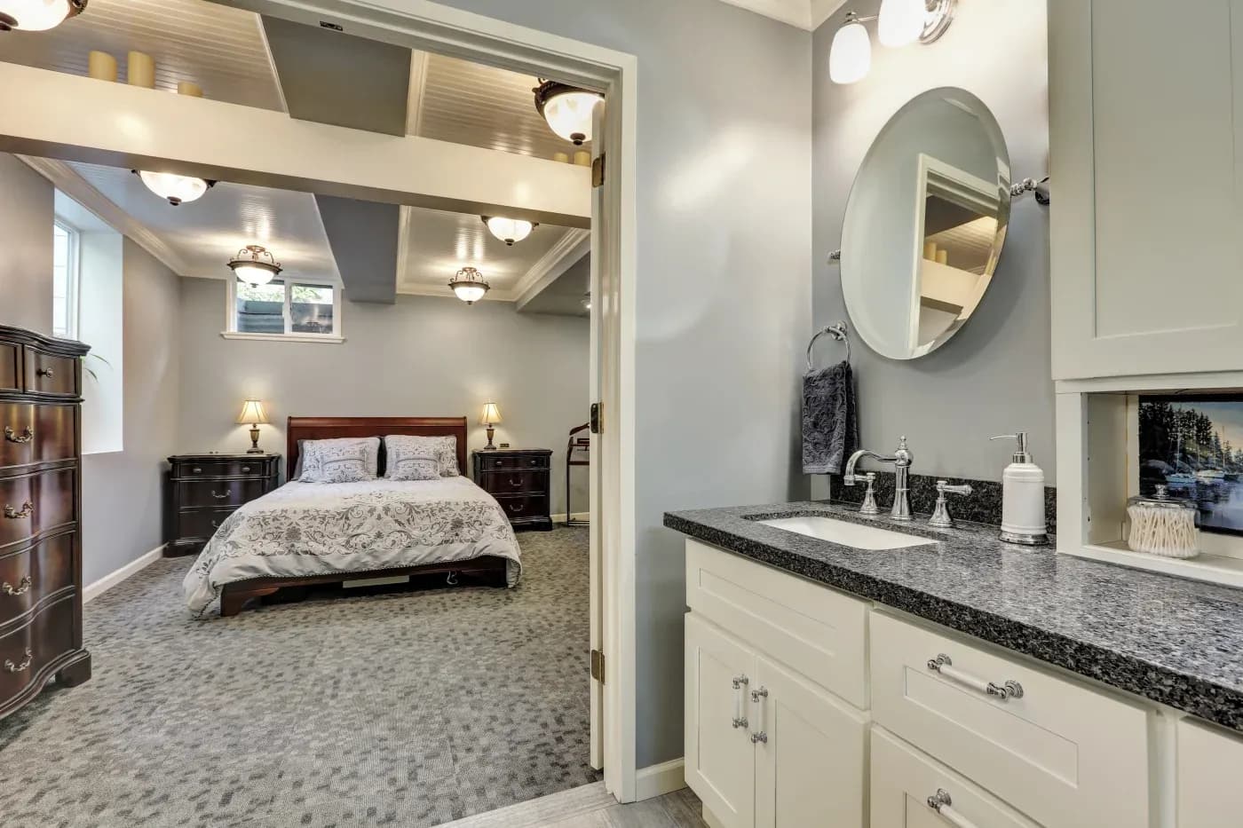 Stylish bedroom with soft gray walls viewed from adjacent bathroom. Features cozy bed, dark wood furniture, carpeted floors, and elegant lighting.
