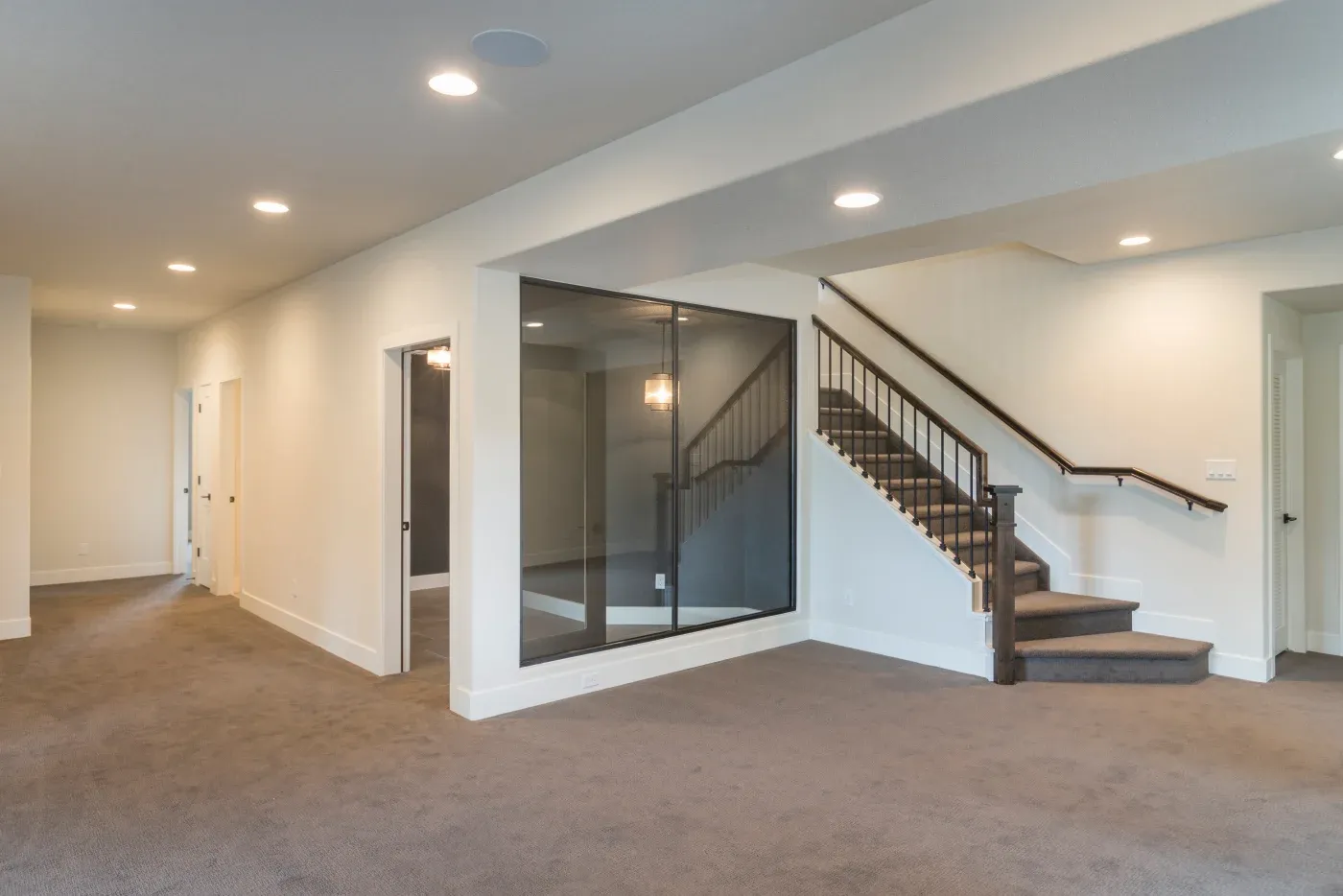 Spacious, modern interior with beige carpet, recessed lighting, and neutral walls. Staircase with iron railing and glass partition adding elegance.