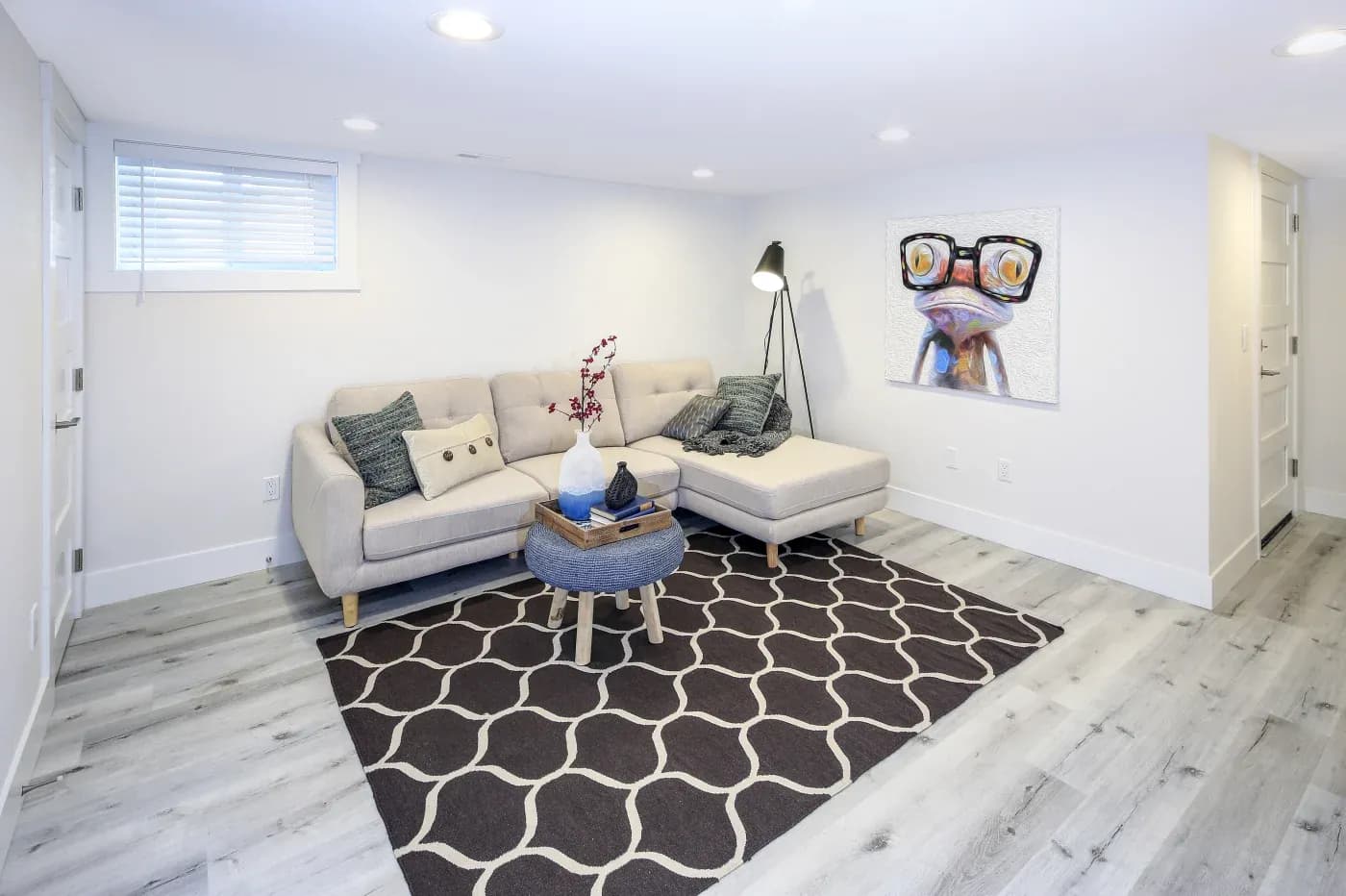 Modern basement living room with light beige sectional sofa, patterned rug, and quirky artwork of a glasses-wearing frog. Bright and cozy ambiance.