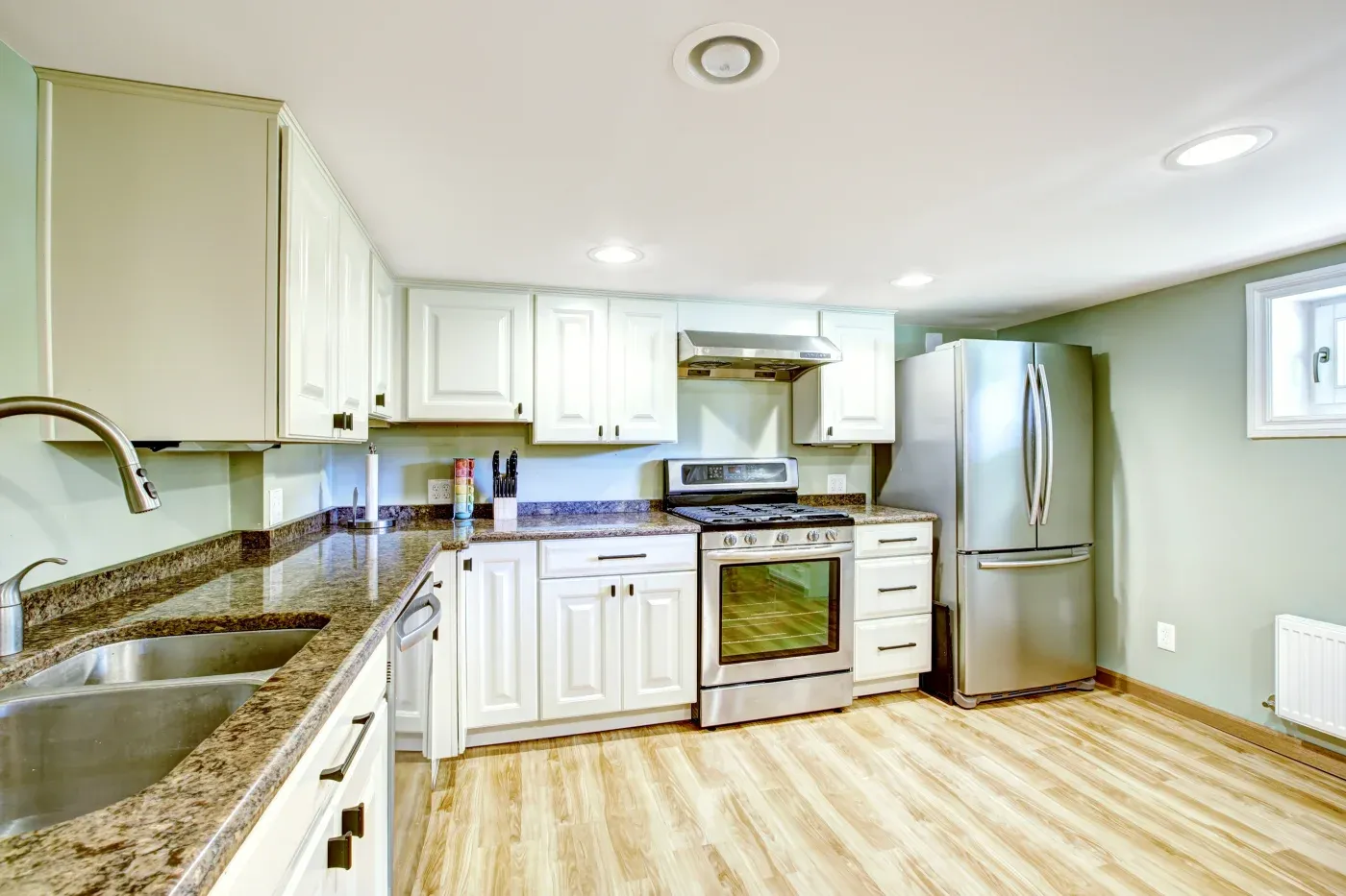 A modern kitchen with white cabinets, stainless steel appliances, and granite countertops. Wooden flooring adds warmth, and recessed lighting creates a bright, clean look.