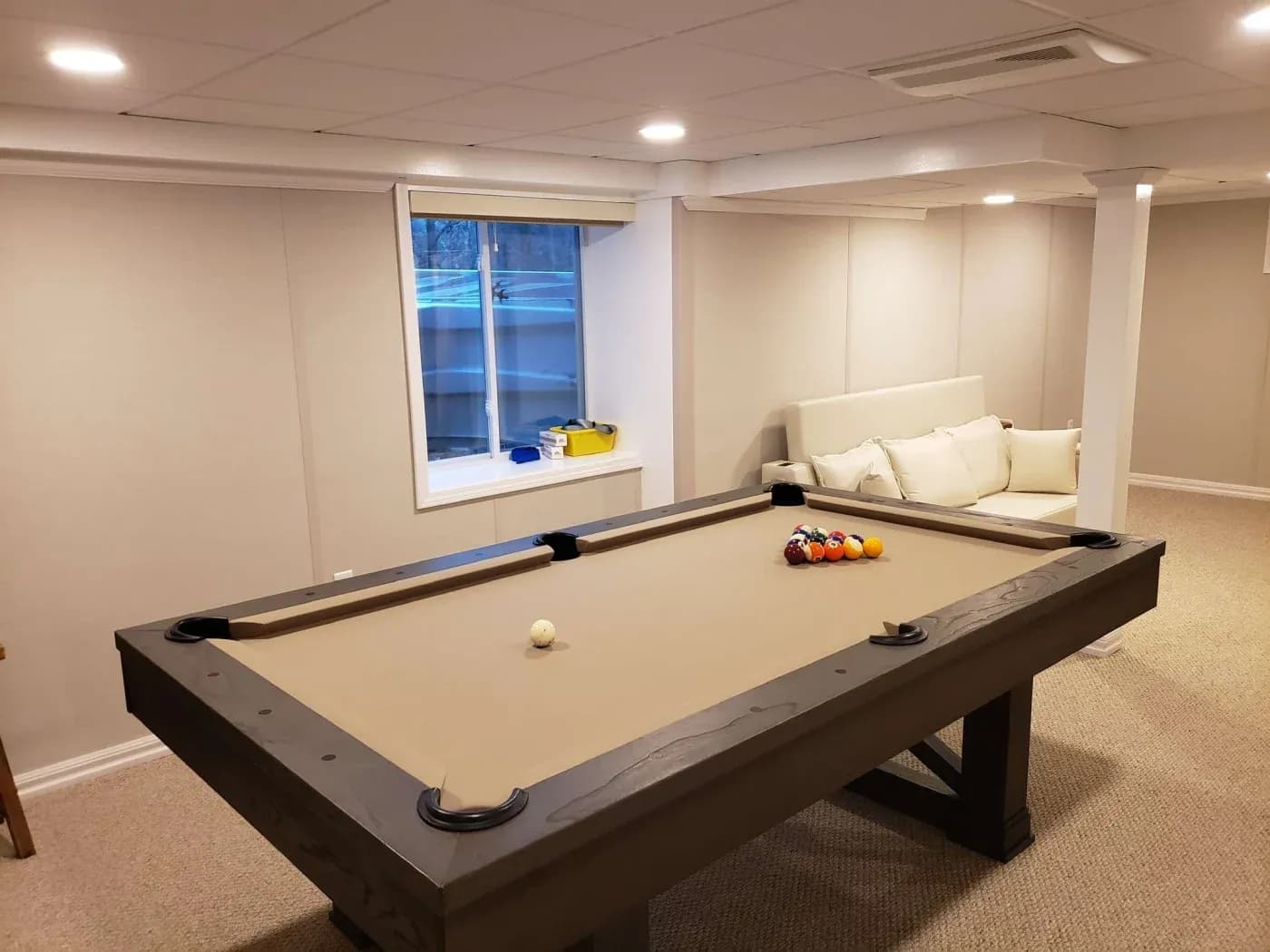 A cozy basement game room featuring a beige pool table with balls set for play. Nearby, a plush white couch sits against a light gray wall, creating a relaxed atmosphere.