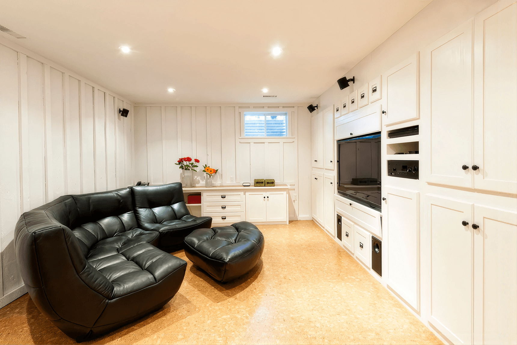 Cozy basement entertainment room with black leather sectional sofa, ottoman, built-in shelves, TV, and white cabinets. Warm lighting adds comfort.