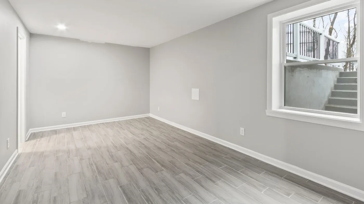 A spacious and empty basement room with light gray walls and wood-like tile flooring. Natural light enters from a window showing outdoor stairs. Minimalist and clean.