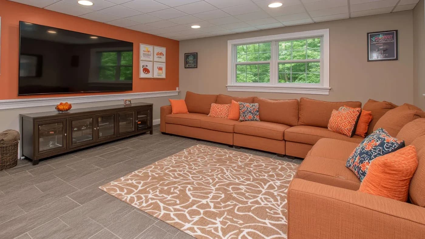 A cozy living room with a large orange sectional sofa adorned with patterned pillows against an orange accent wall. A flat-screen TV hangs above a dark wood cabinet. A beige and white patterned rug covers the tile floor. Large window provides natural light, creating a welcoming atmosphere.