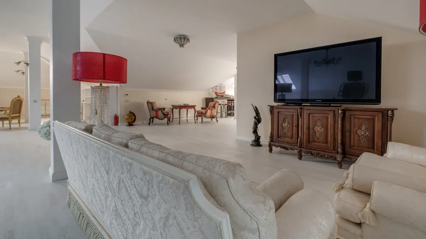 Spacious living room with elegant decor, featuring a large TV on a carved wooden cabinet, luxury cream sofas, a red lamp, and a bright, airy ambiance.