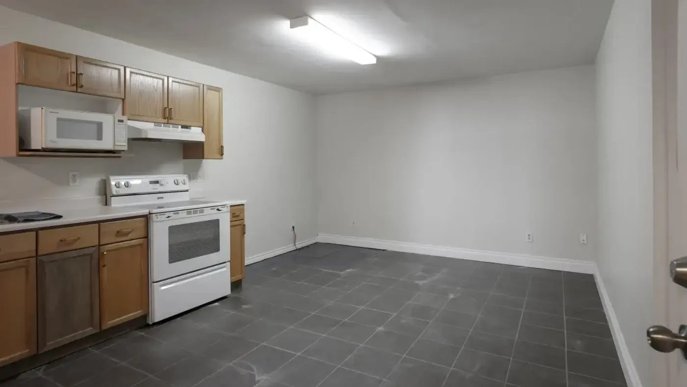 A minimalist kitchen with light wooden cabinets, a white stove, and microwave. It has gray tile flooring, white walls, and a bright overhead light.
