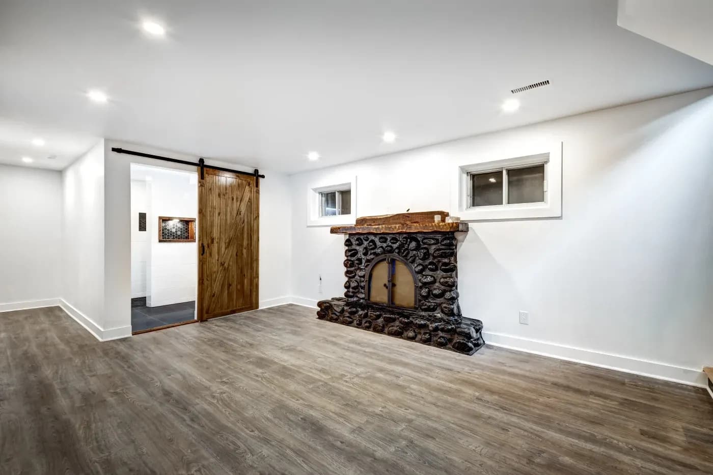 Spacious room with wood flooring, a rustic stone fireplace, and a wooden sliding barn door. White walls, ceiling lights, and two small windows enhance brightness.