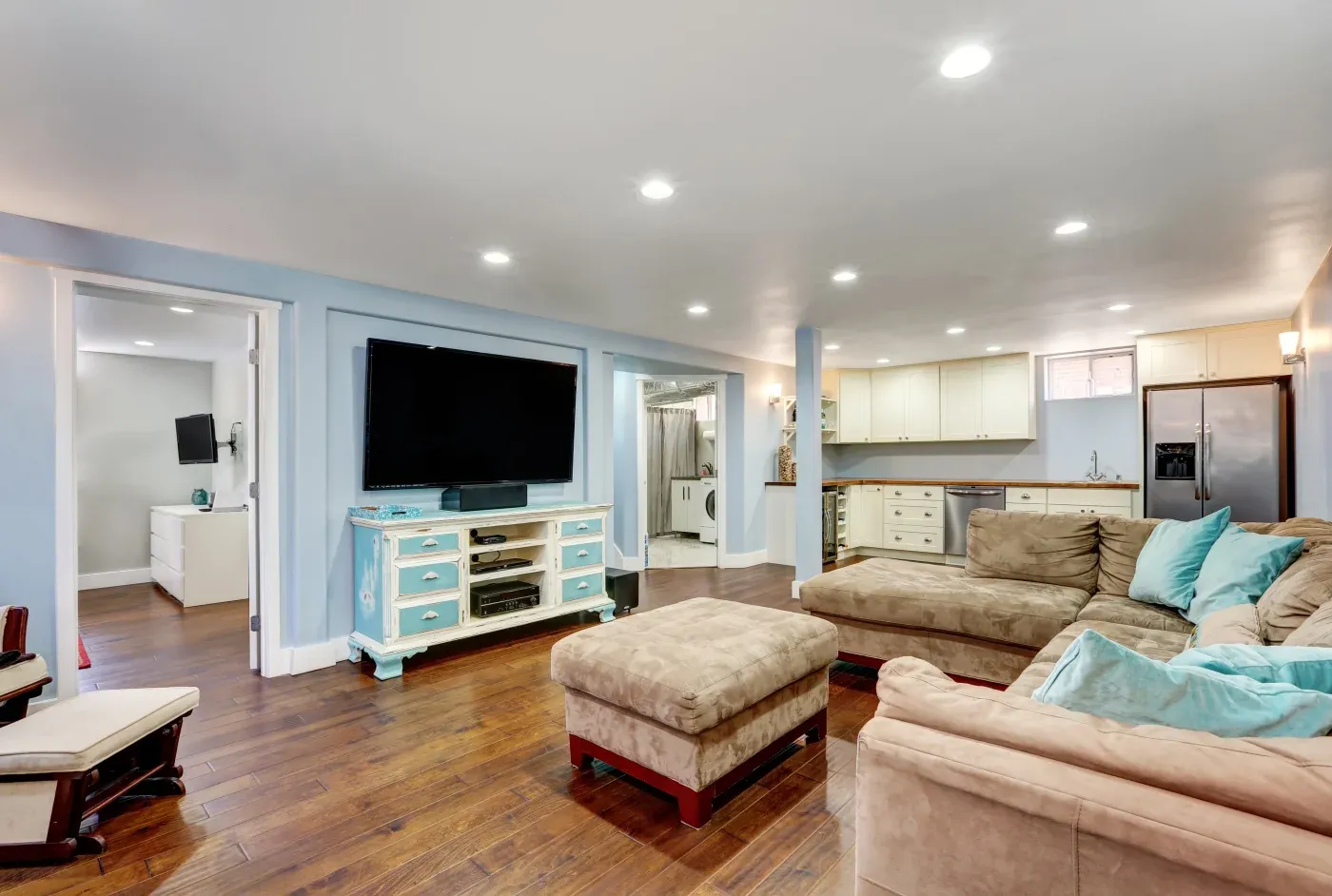 Spacious basement living area with blue walls, featuring a large sectional sofa and ottoman. A flat-screen TV is on a painted console. Kitchen area with stainless steel appliances is visible in the background, creating a cozy and inviting atmosphere.