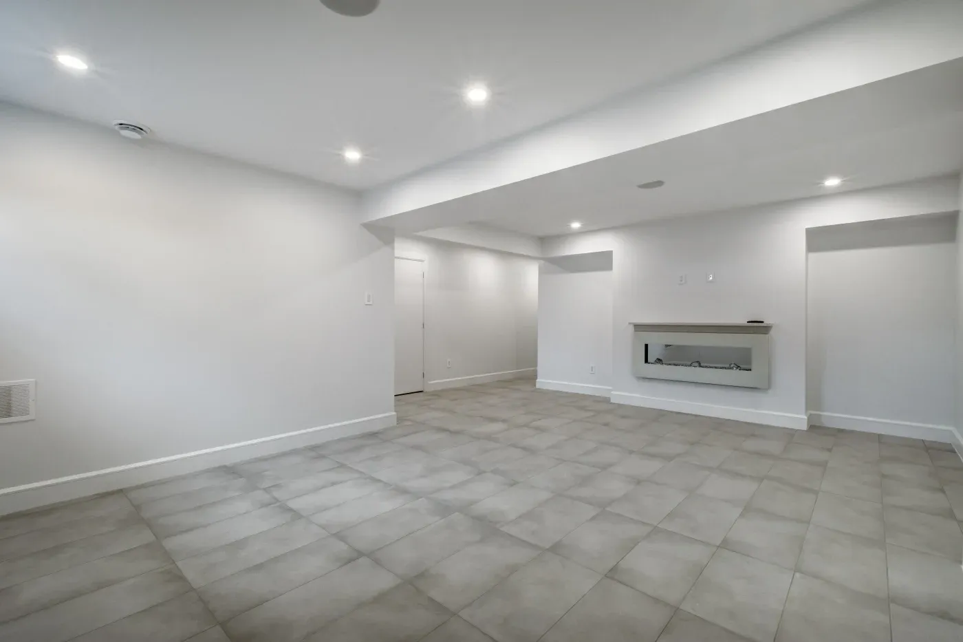 Spacious, modern basement with light gray tiles, white walls, and recessed lighting. Features a wall-mounted electric fireplace and clean lines.