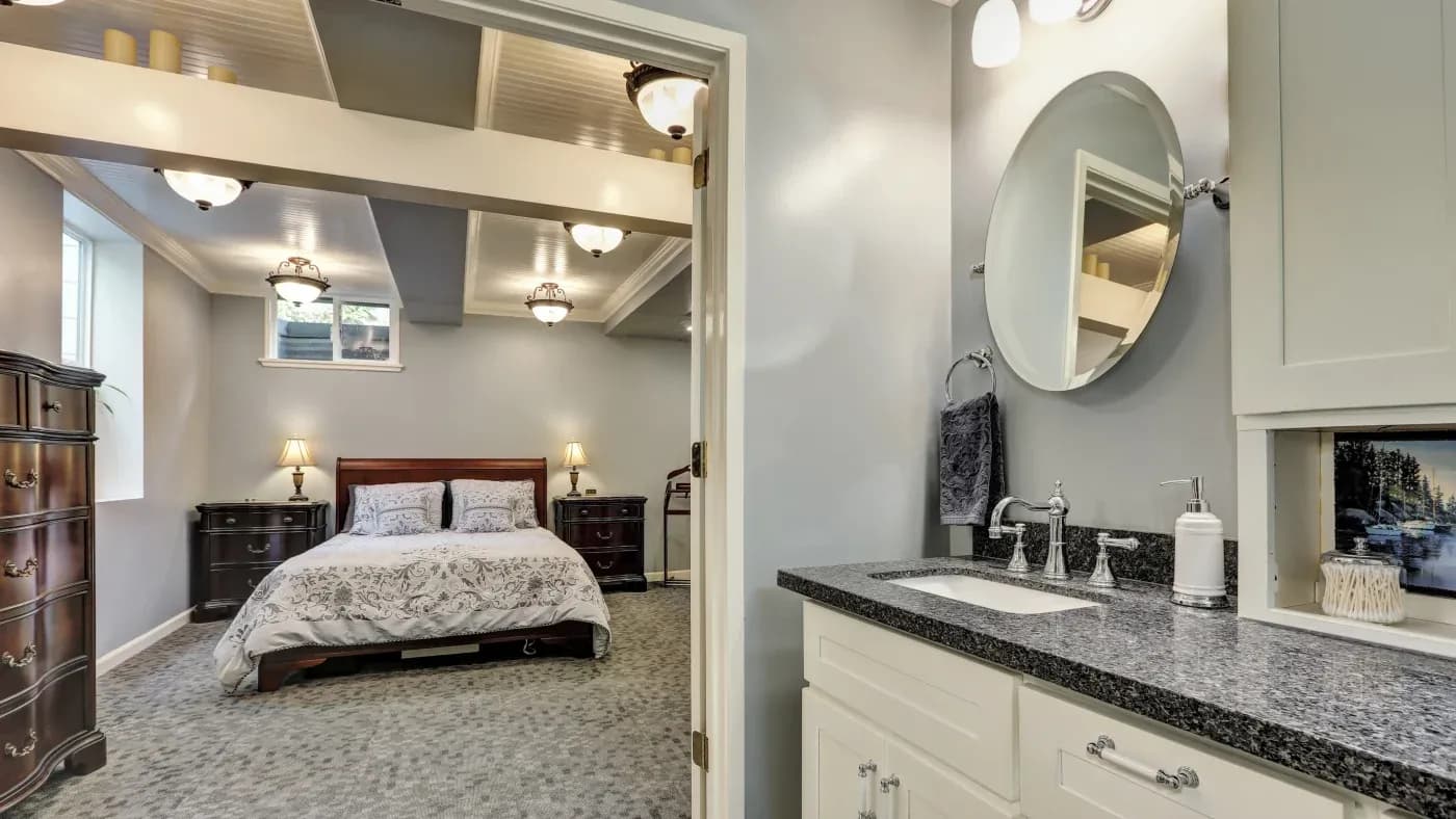 A cozy bedroom with gray walls features a bed with white patterned bedding. Two wooden nightstands with lamps sit beside it. A doorway leads to a bathroom with a granite countertop and round mirror, conveying a calm, elegant ambiance.