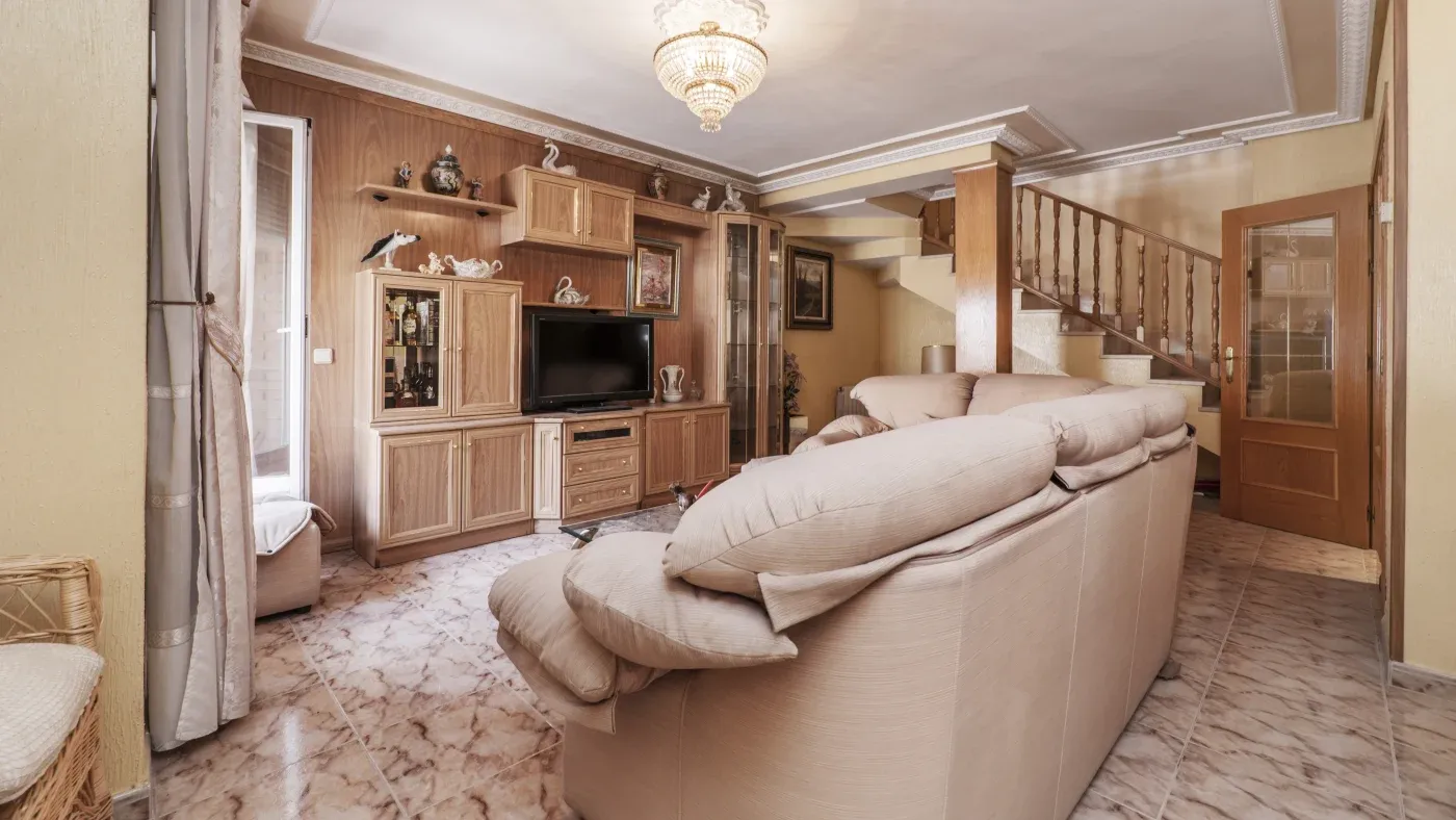 Cozy living room with beige sofas facing a wooden entertainment center and flat-screen TV. Marble flooring, elegant chandelier, and a staircase create a warm, inviting atmosphere.
