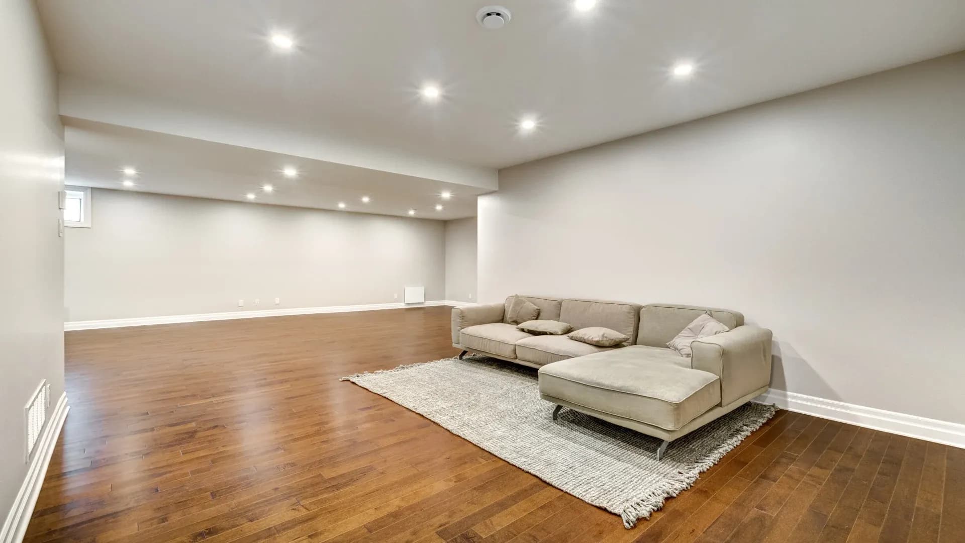 Spacious basement with wooden floors, beige walls, and recessed lighting. A gray sectional sofa sits on a textured rug, creating a cozy ambiance.
